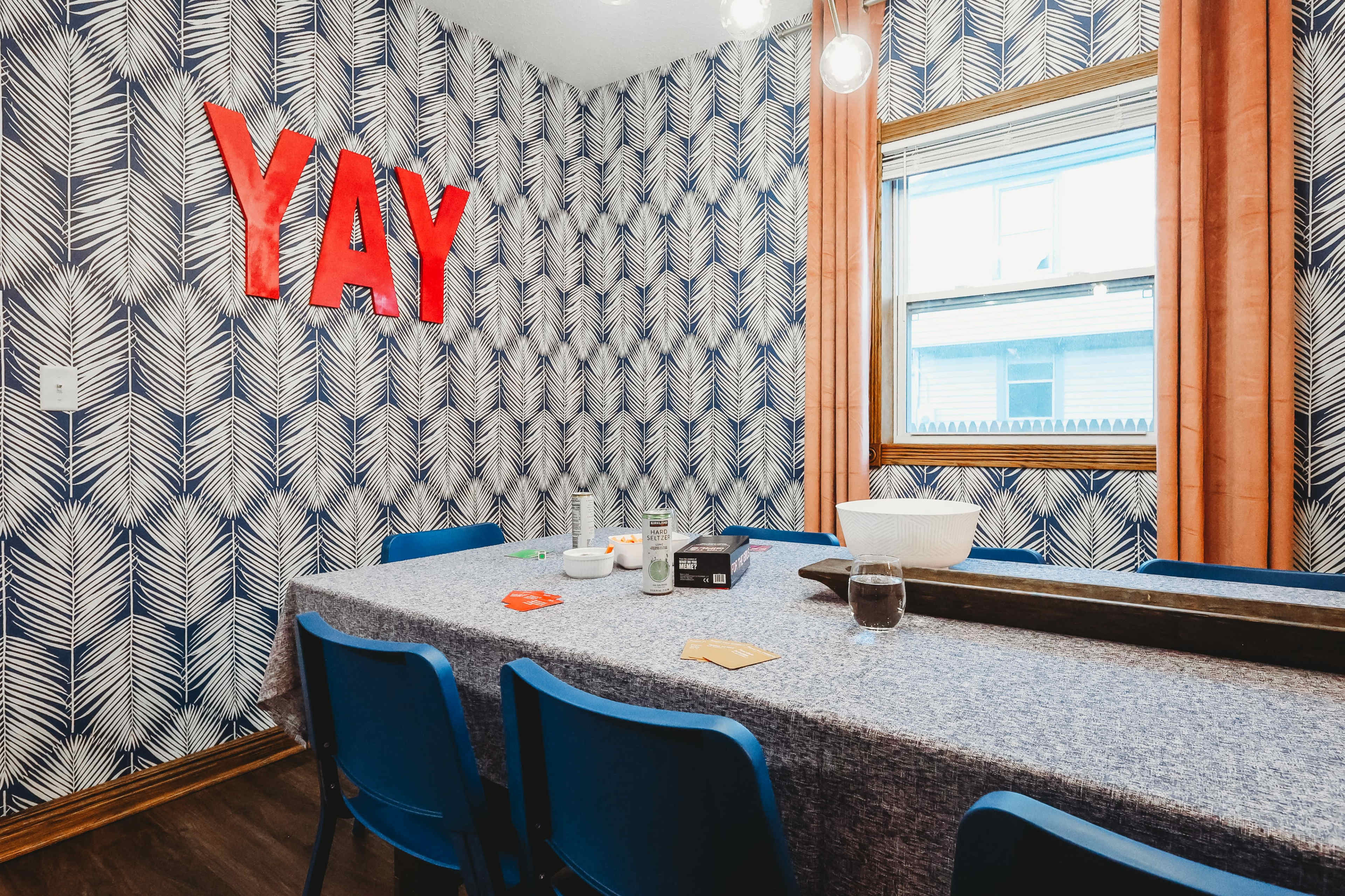 A dining room features a blue table covered with a cloth, surrounded by blue chairs, with a wall decorated in a leafy pattern and a bright red "YAY" sign.