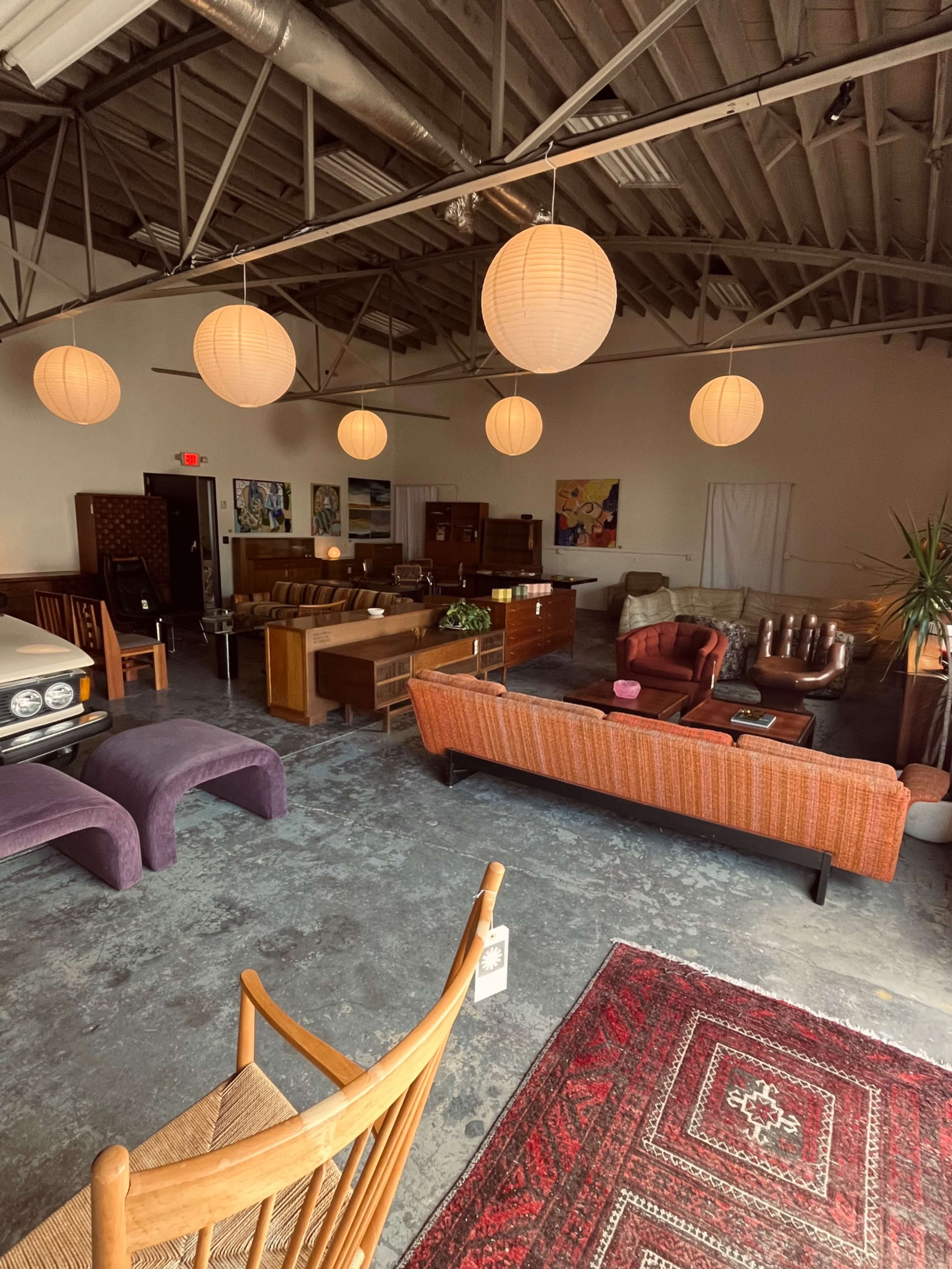 The image shows a spacious interior with various vintage-furniture pieces, including a large orange couch, a wooden rocking chair, and decorative paper lanterns hanging from the ceiling.