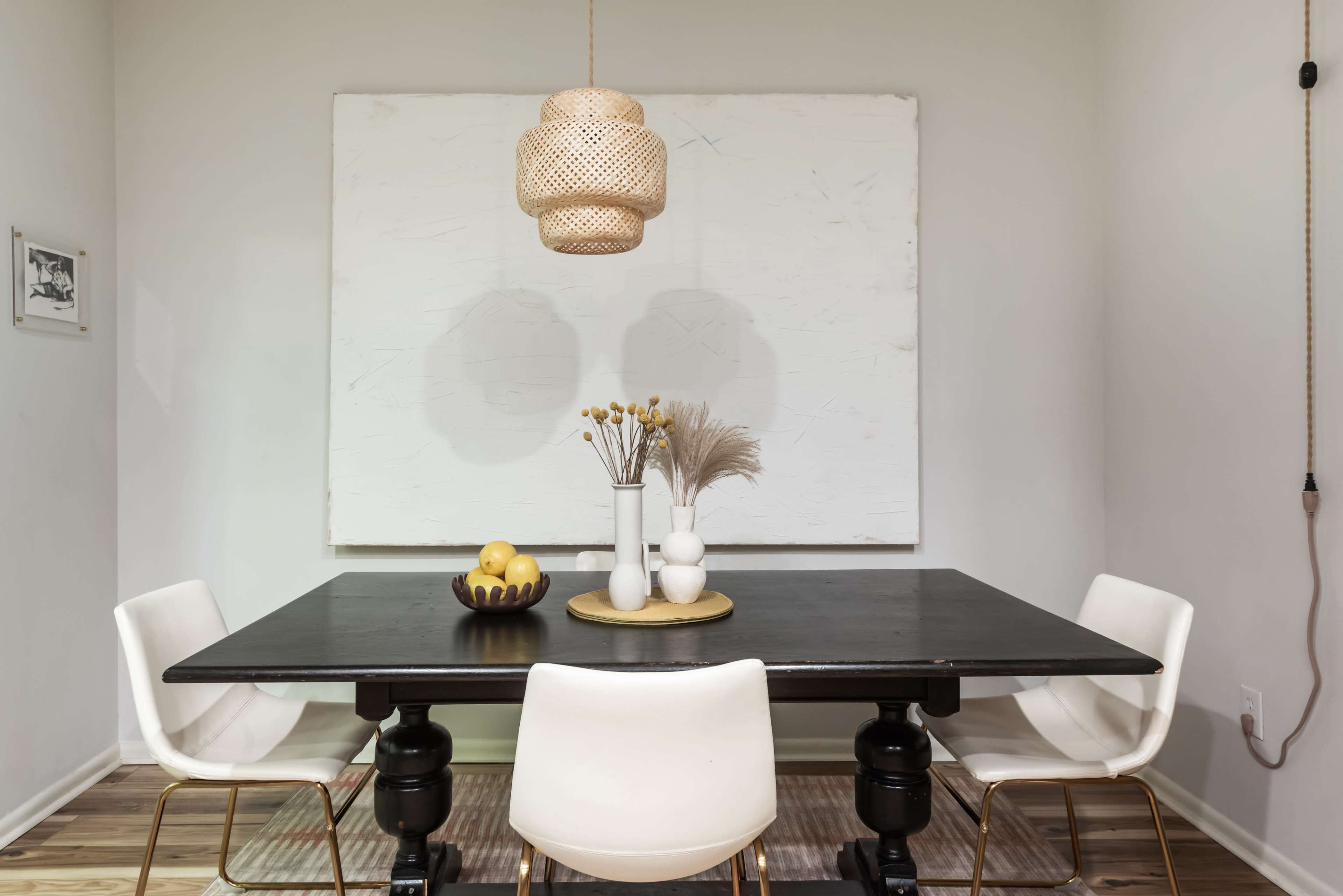 A modern dining area features a black table surrounded by white chairs, with a large abstract painting on the wall and a woven pendant light overhead.