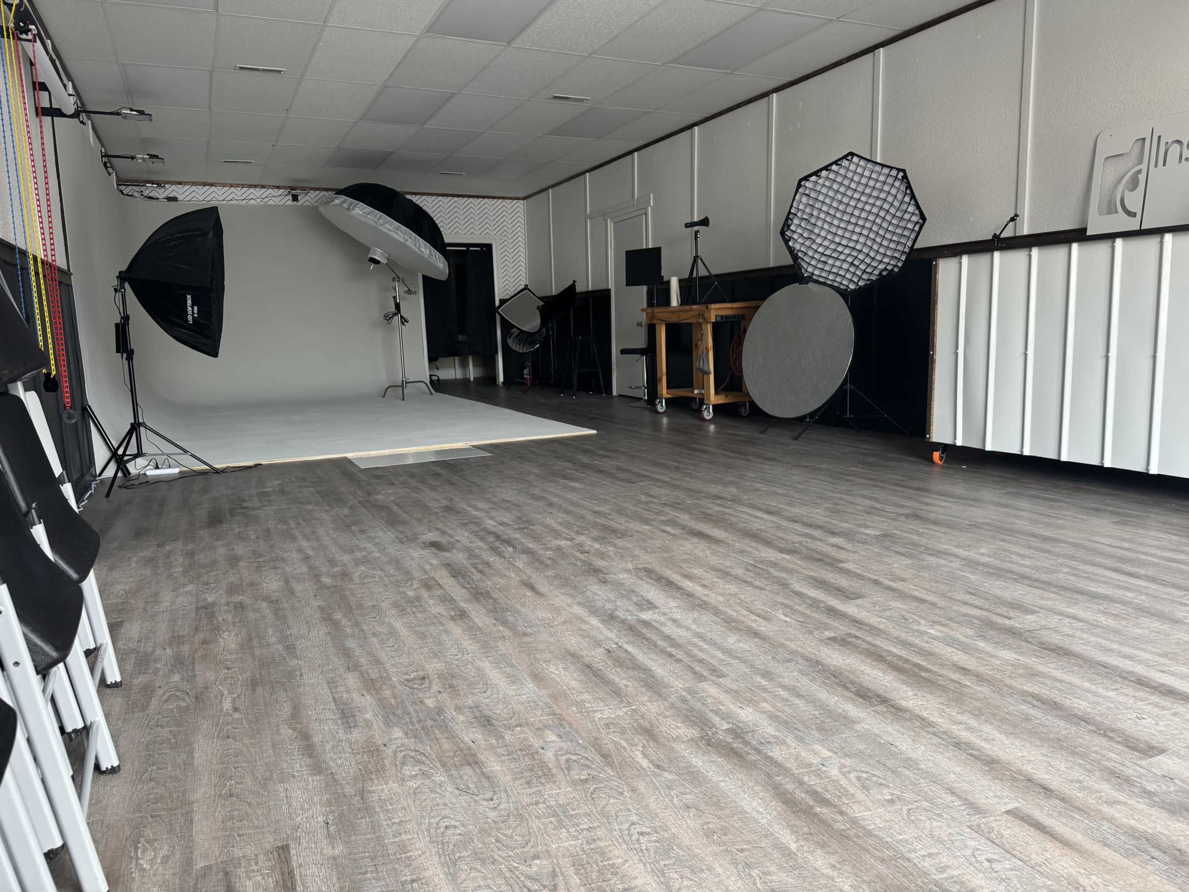 The image shows a spacious photography studio featuring a light gray backdrop, various lighting equipment, and a wooden table against a white wall.