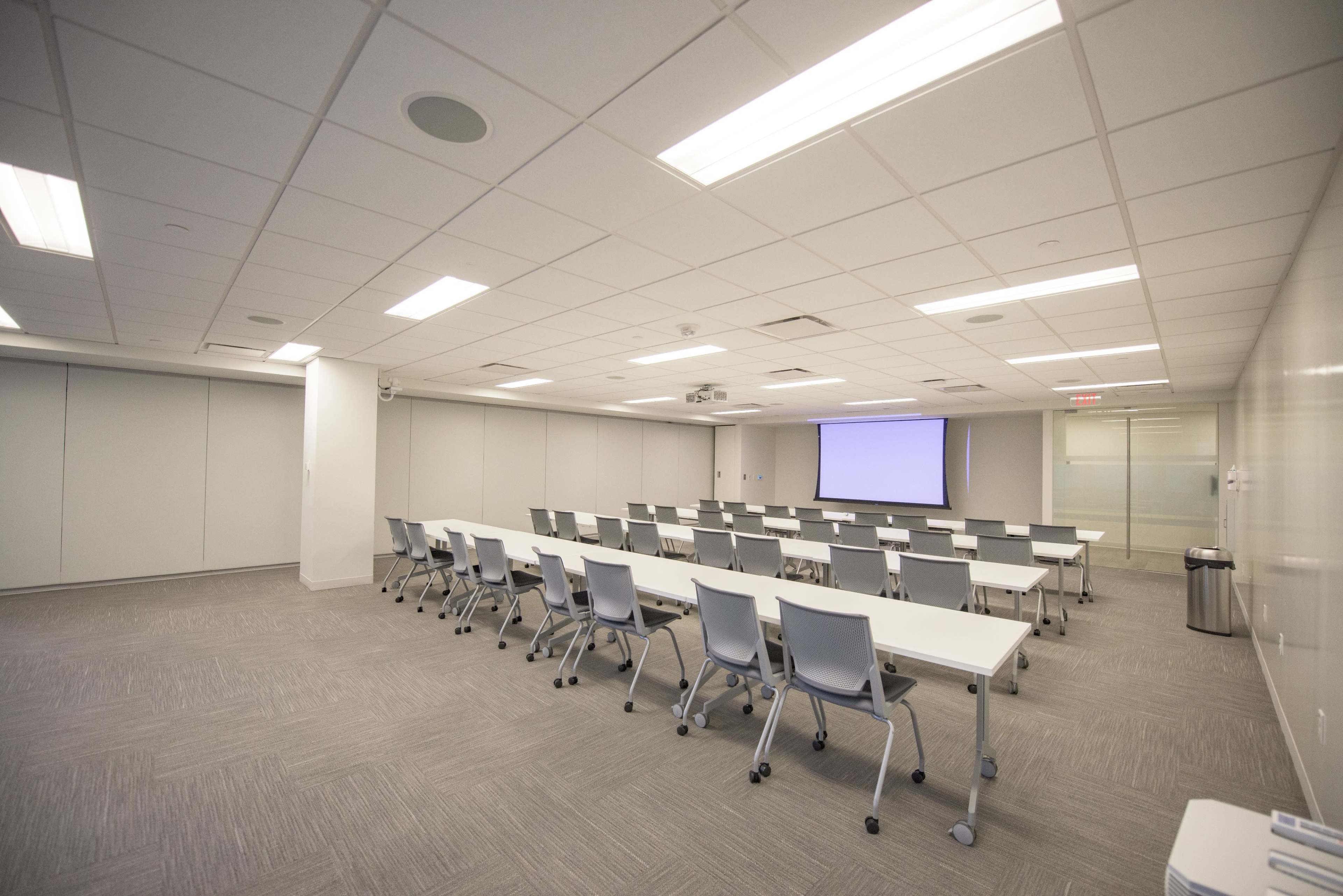 A conference room with rows of tables and chairs, equipped with a projector screen at one end.