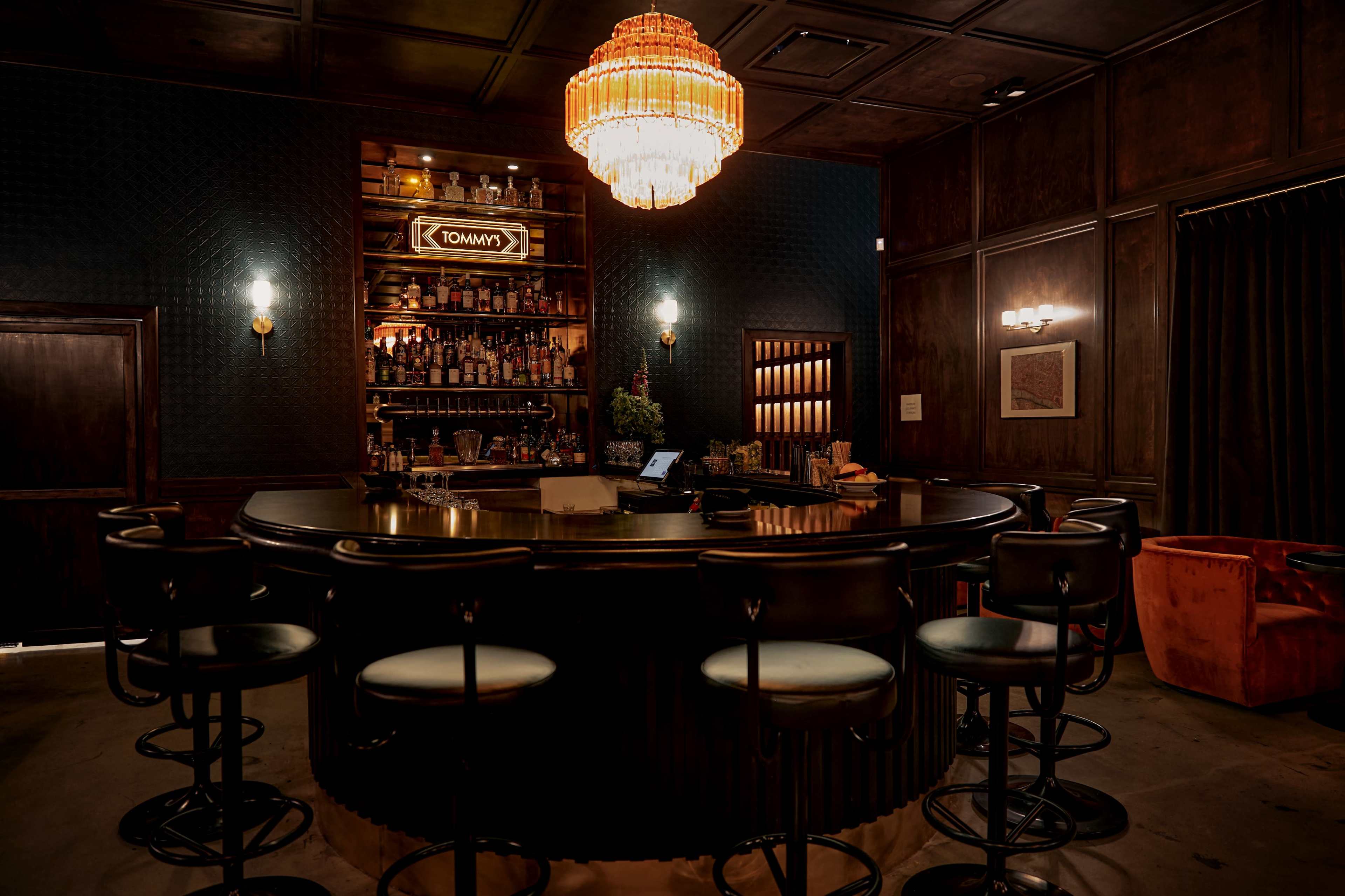 A dimly lit bar with a circular wooden counter, high stools, and a chandelier above, surrounded by dark wooden walls and shelves filled with bottles.