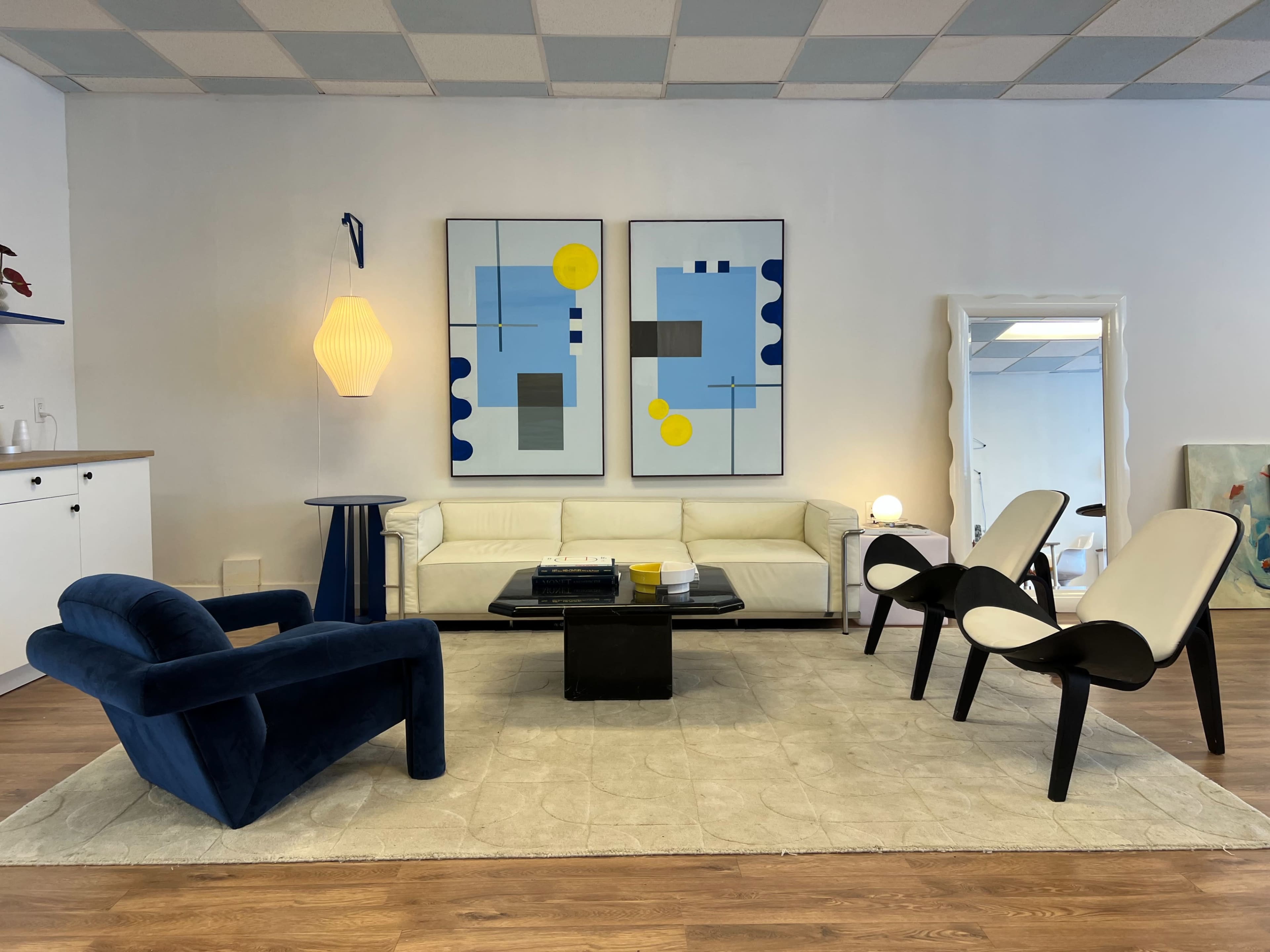 A modern living room with a white sofa, black coffee table, and two stylized chairs arranged on a light-colored rug, featuring abstract artwork on the walls.
