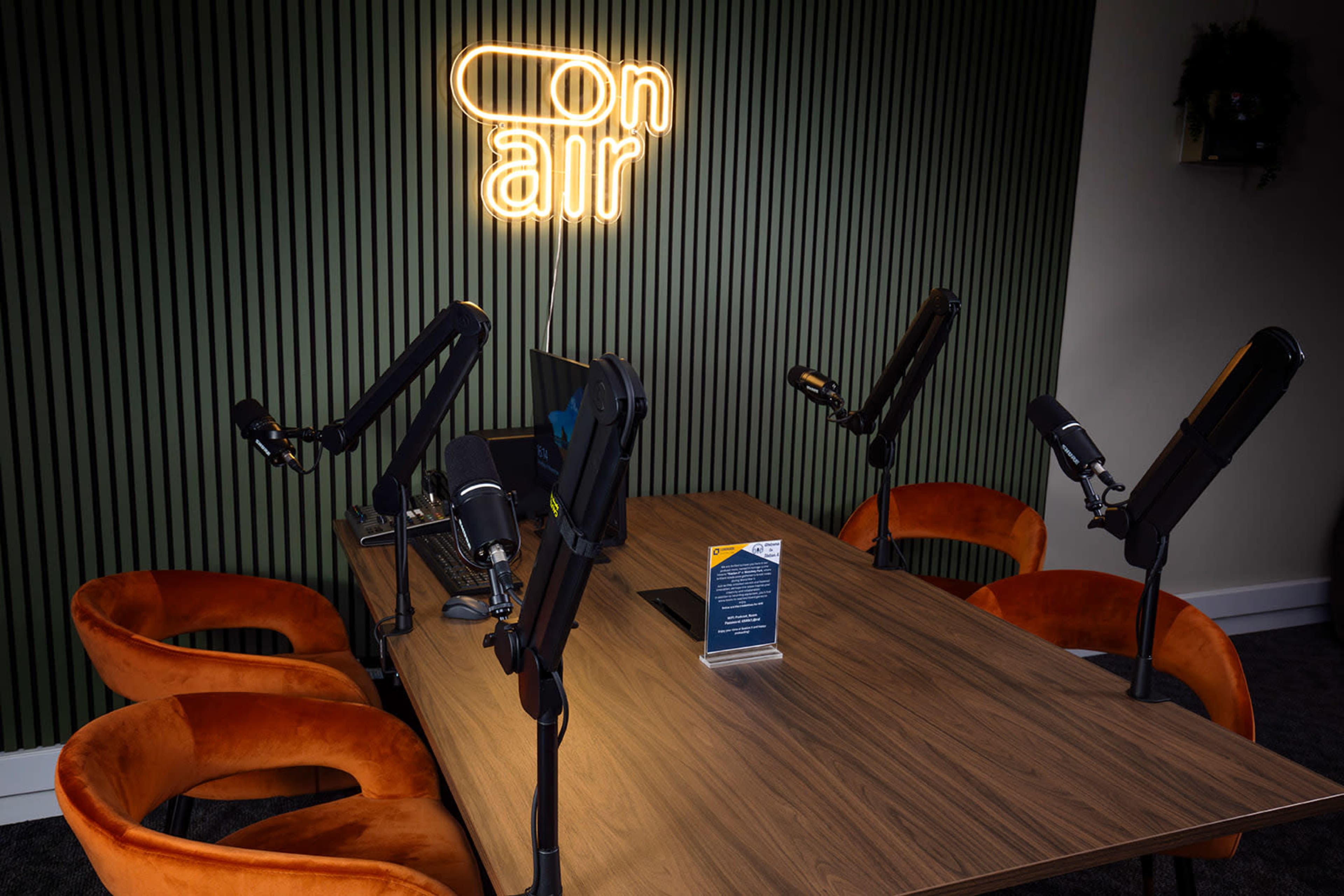 A modern podcast studio features a wooden table with four microphones and several orange chairs, illuminated by a neon "on air" sign on the wall.