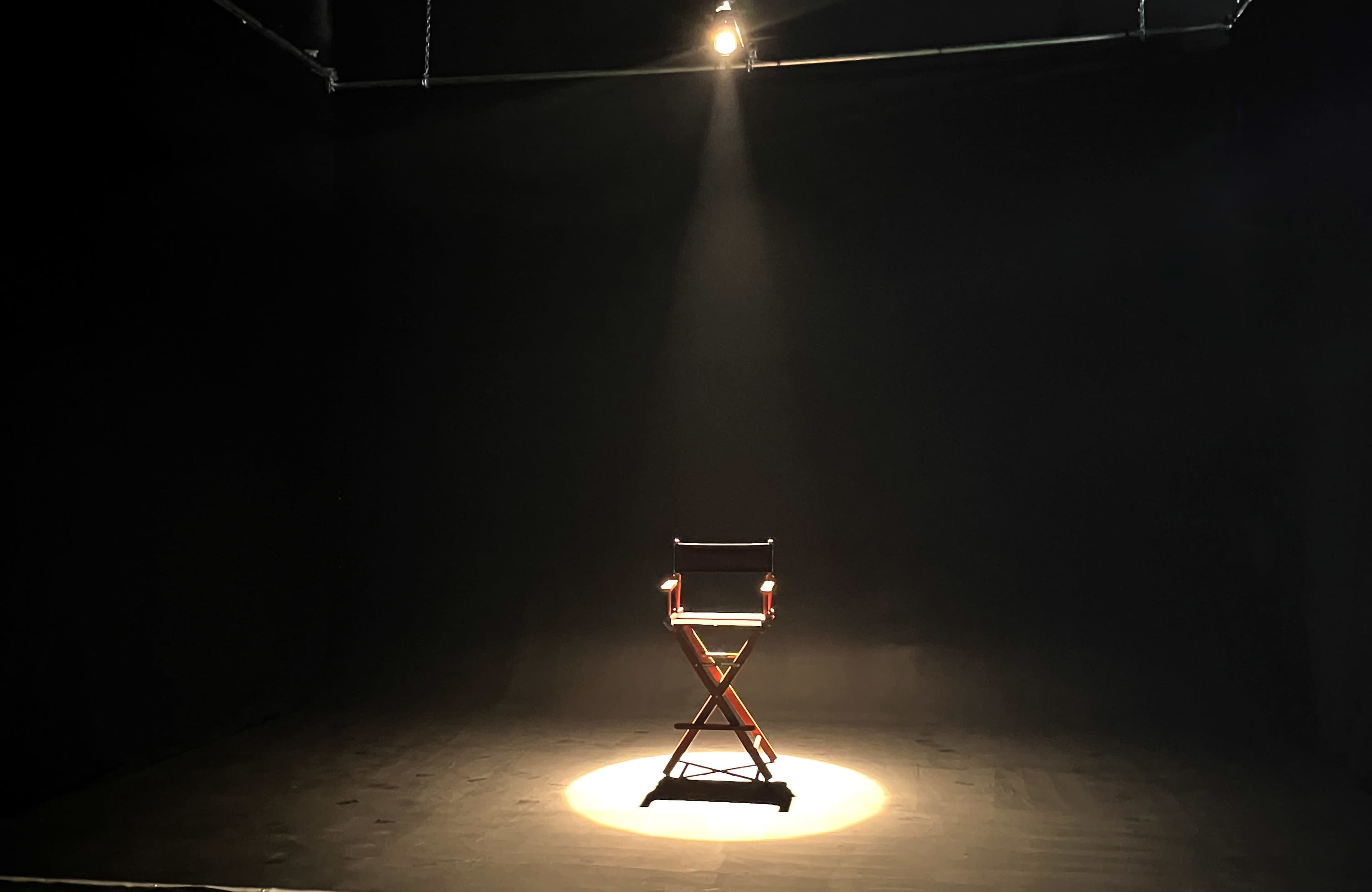 A wooden director's chair is illuminated by a spotlight on a dark stage.