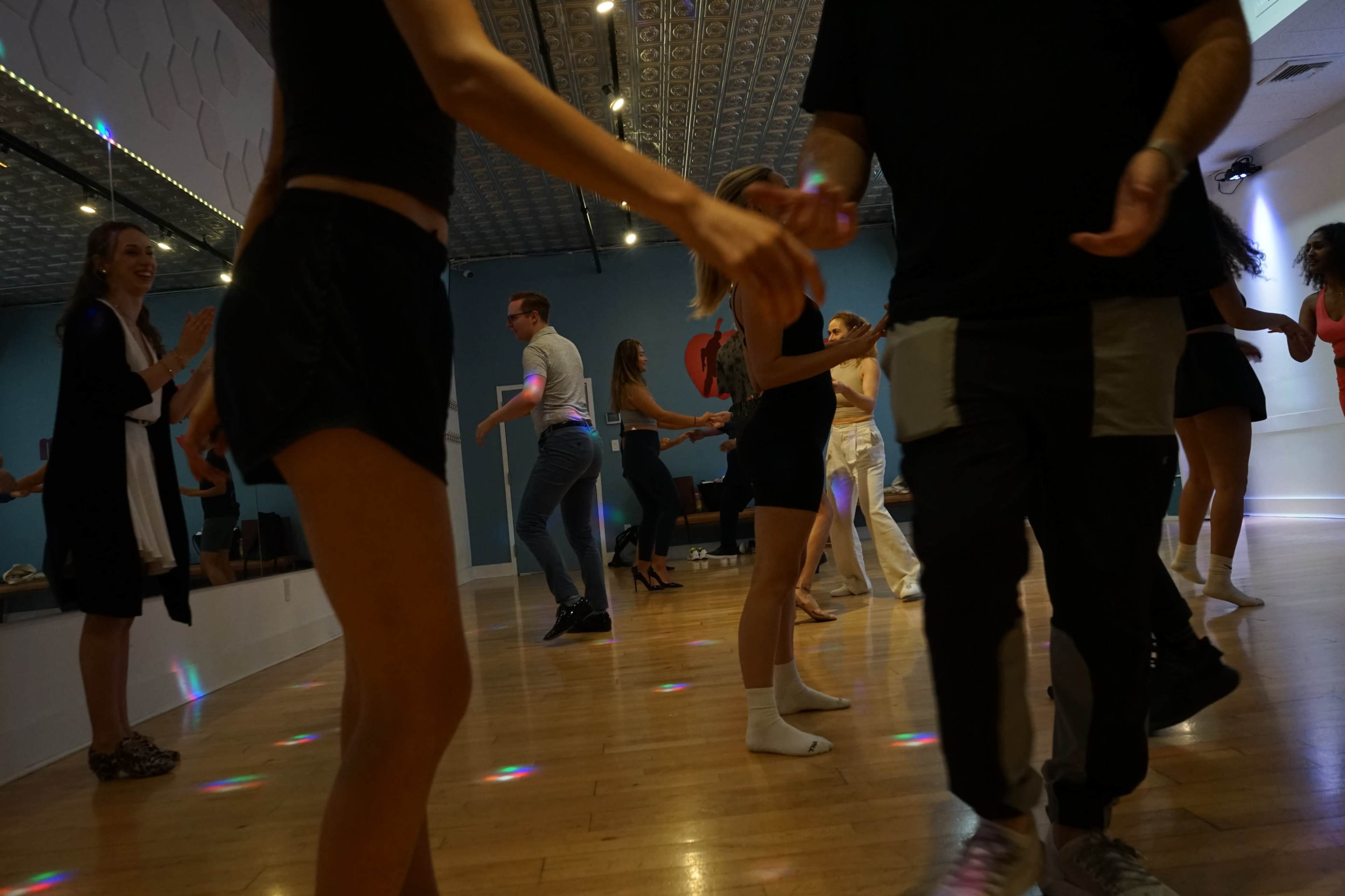 A group of people dance together in a well-lit studio featuring a mirrored wall and colorful lights on the wooden floor.