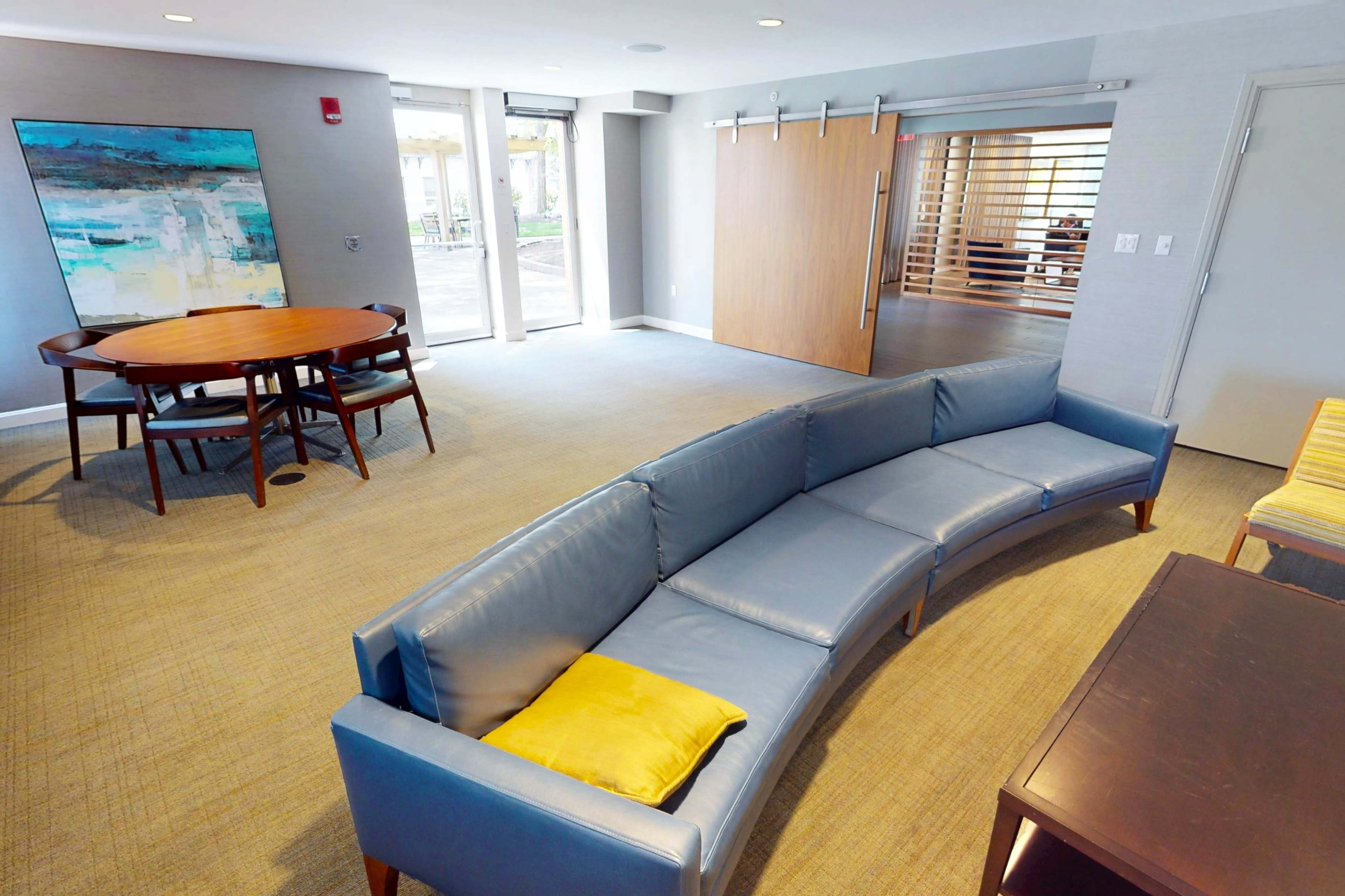 The image shows a modern lounge area featuring a curved blue sofa, a round wooden dining table, and a large abstract painting on the wall.
