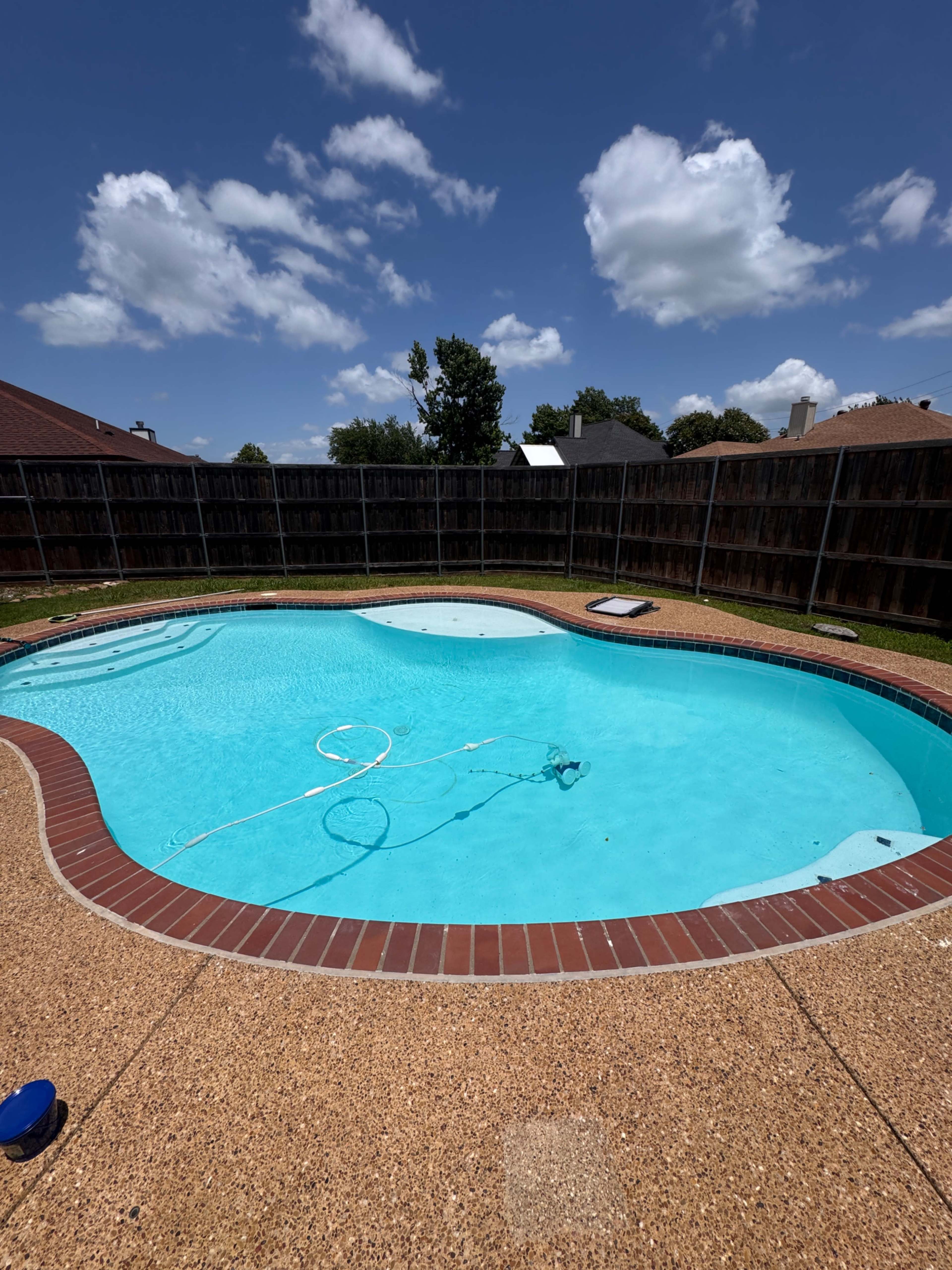 A clear blue swimming pool is surrounded by a brown brick patio and a wooden fence under a partly cloudy sky.