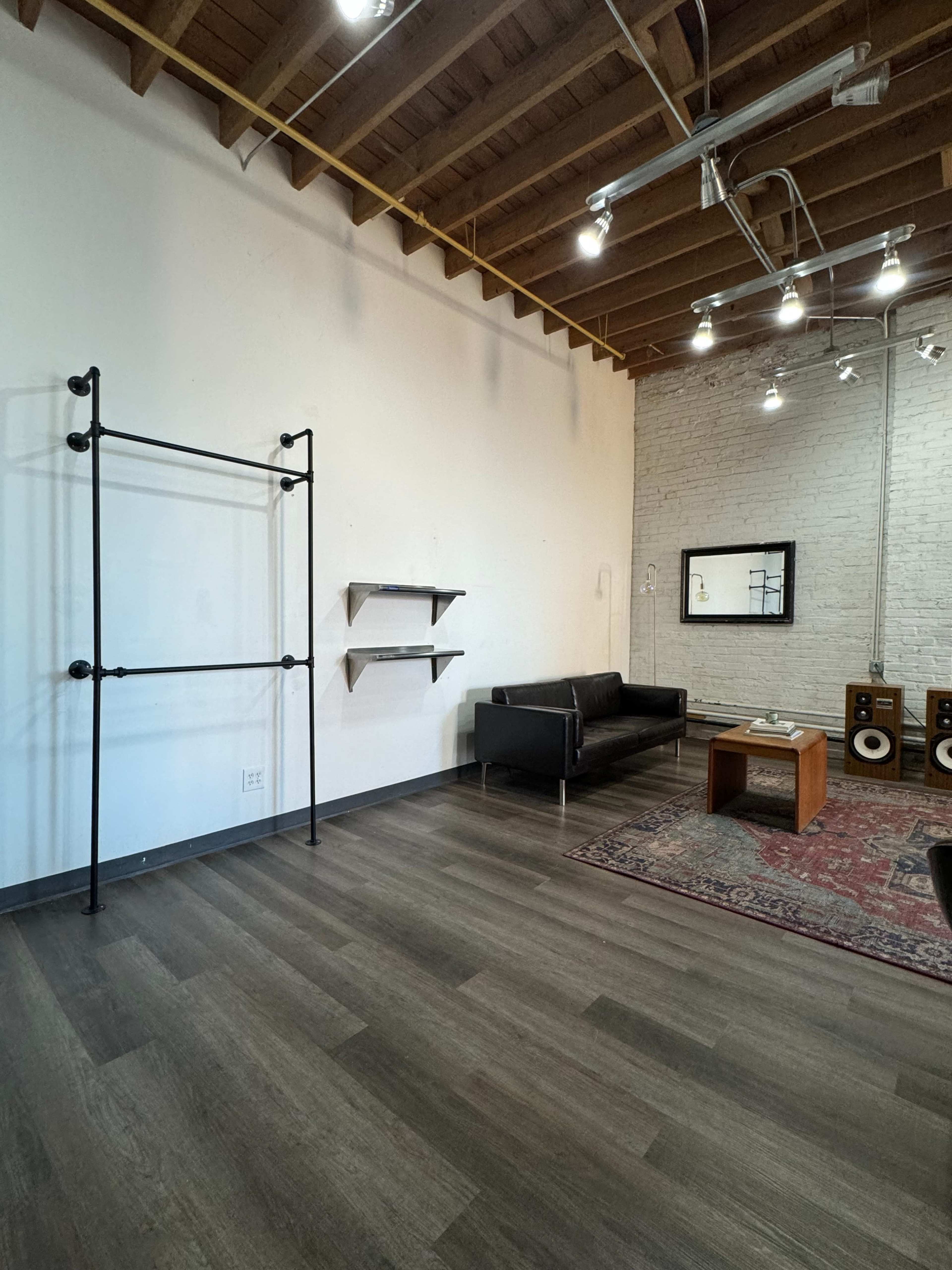 The image shows a spacious interior featuring a black couch, a coffee table, wooden flooring, and a couple of shelves attached to one wall, with exposed brick and wooden beams overhead.