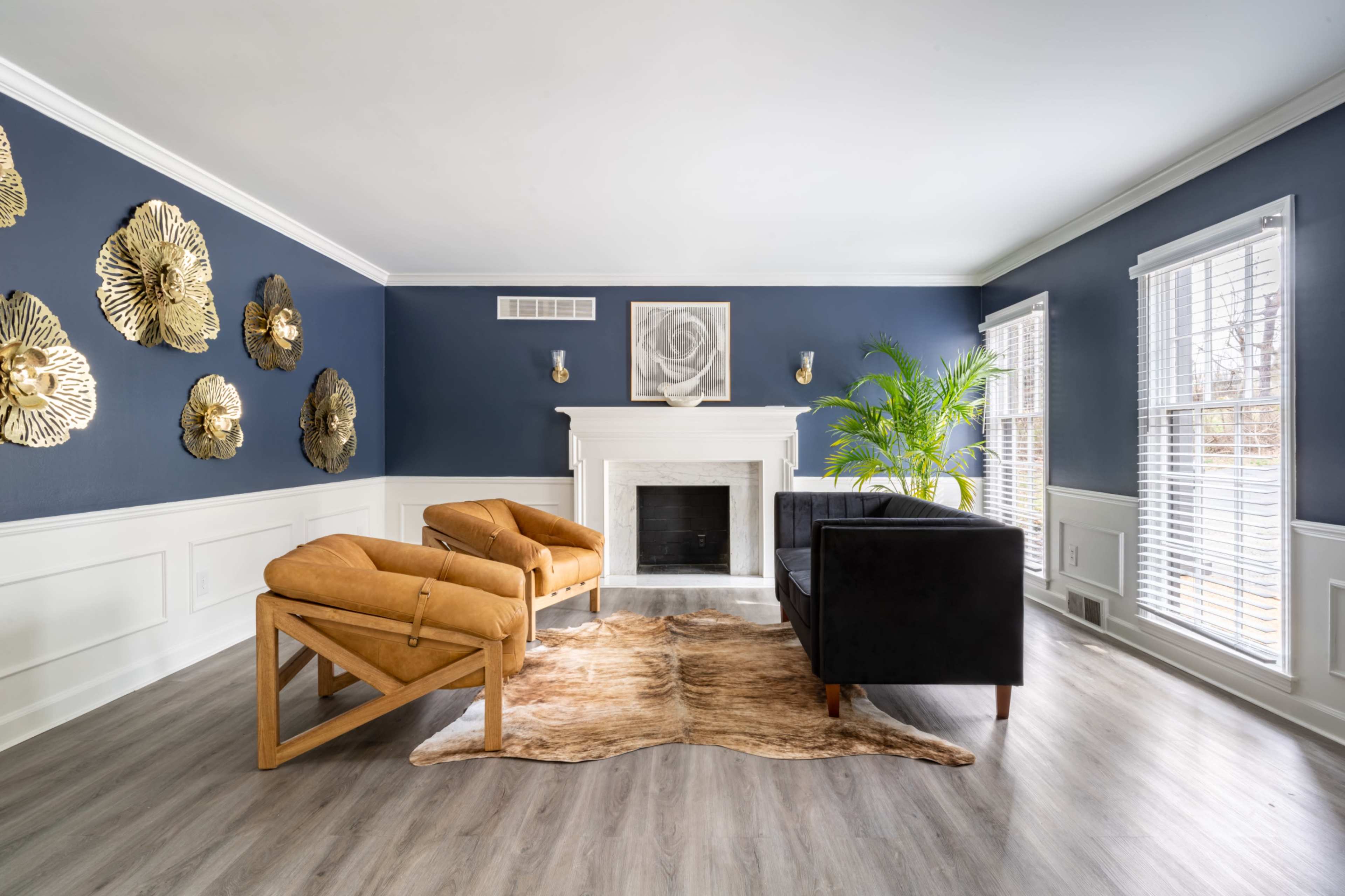 The room features a fireplace with a white mantel, two tan armchairs, a black sofa, a rug, and decorative wall art made of gold-colored flowers against dark blue walls.