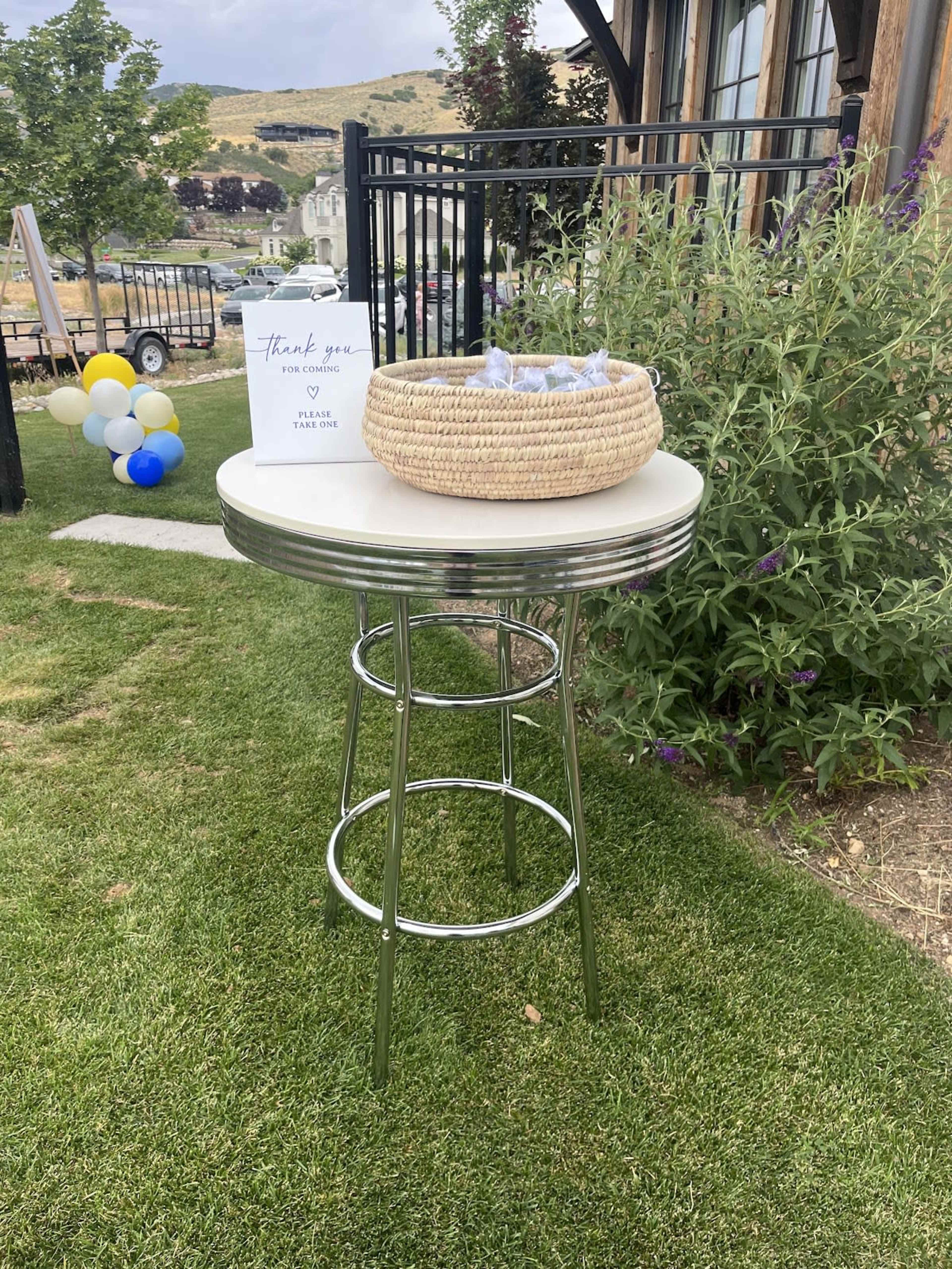 A small round table with a chrome base is set on green grass, holding a woven basket and a sign that expresses gratitude.