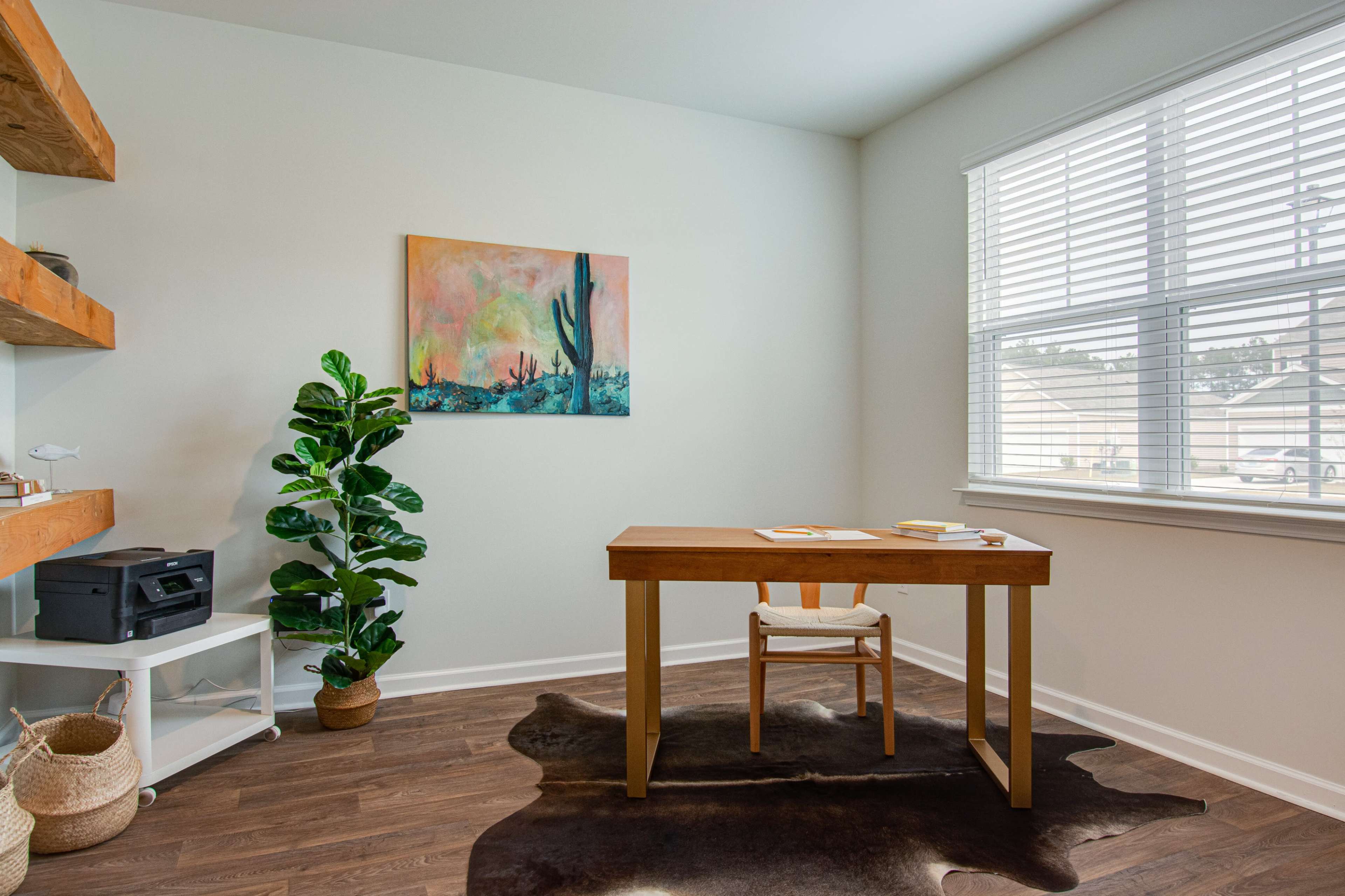A minimalist office features a wooden desk, a chair, a printer, and a potted plant under a colorful wall painting, illuminated by natural light from a window.