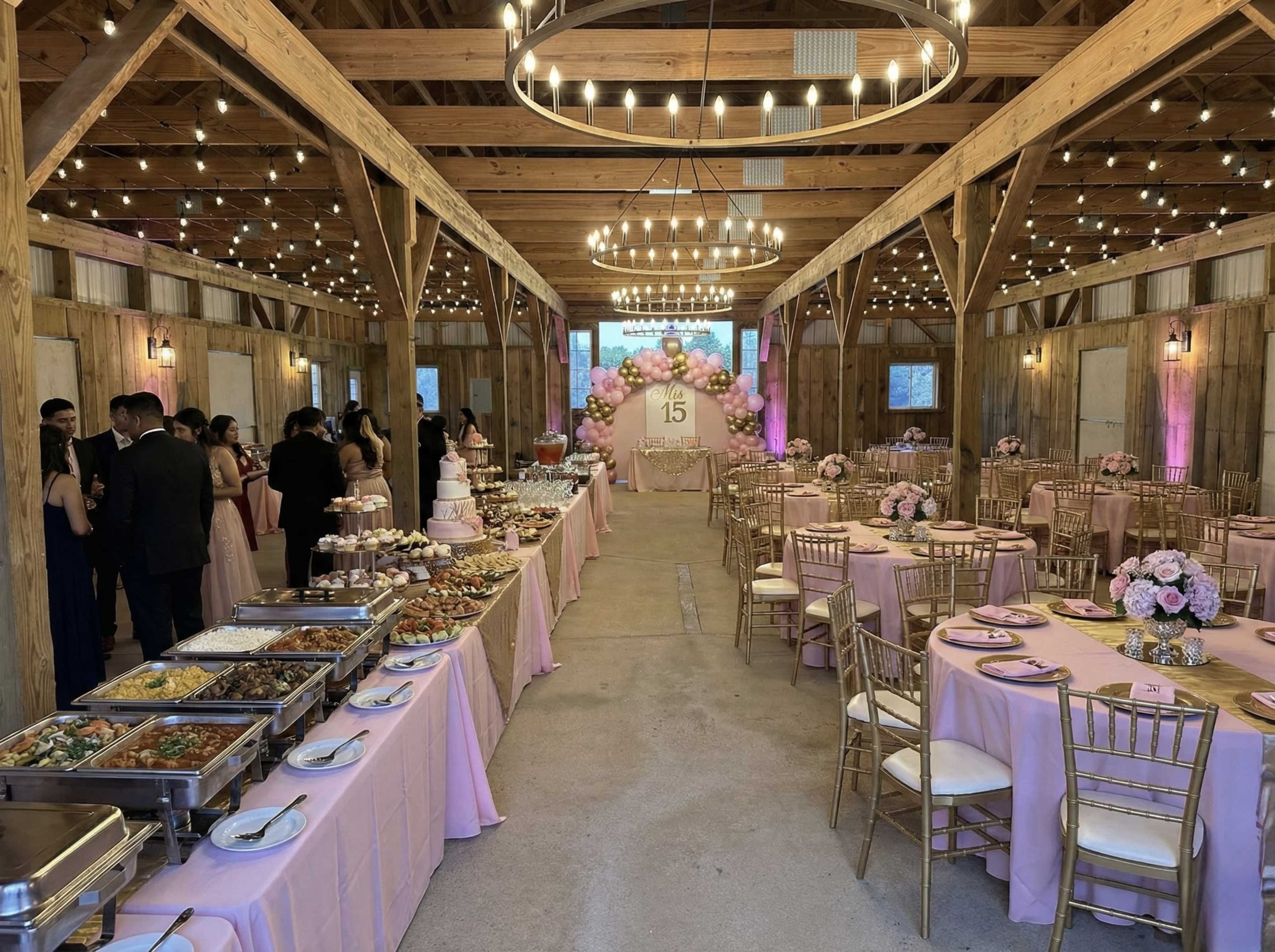 The image shows a decorated event space set up for a celebration, featuring tables with pink tablecloths, floral centerpieces, a buffet line, and a backdrop of balloons and a number "15."