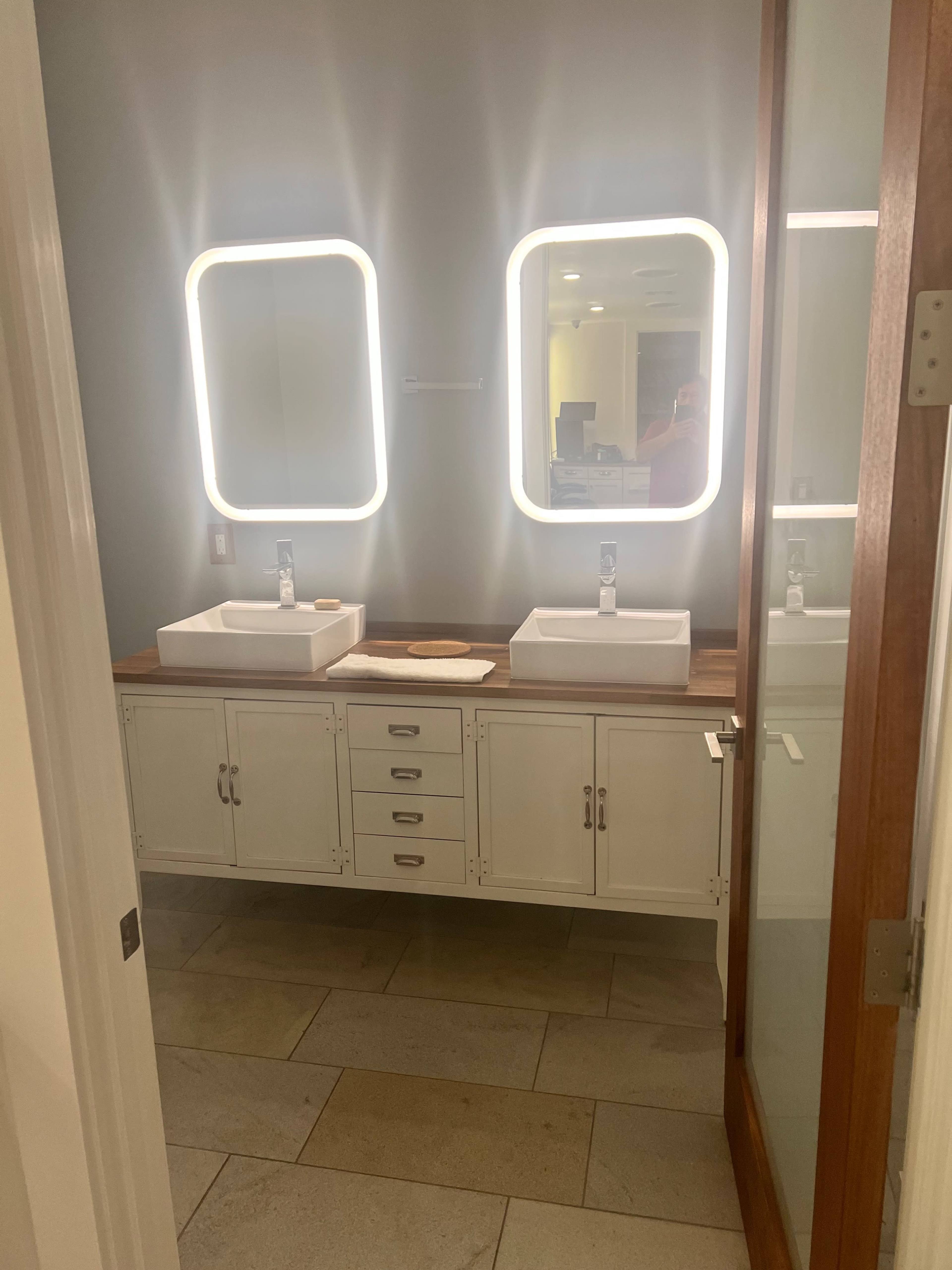 The image shows a modern bathroom with two rectangular sinks, illuminated mirrors, and wooden cabinetry on a tiled floor.