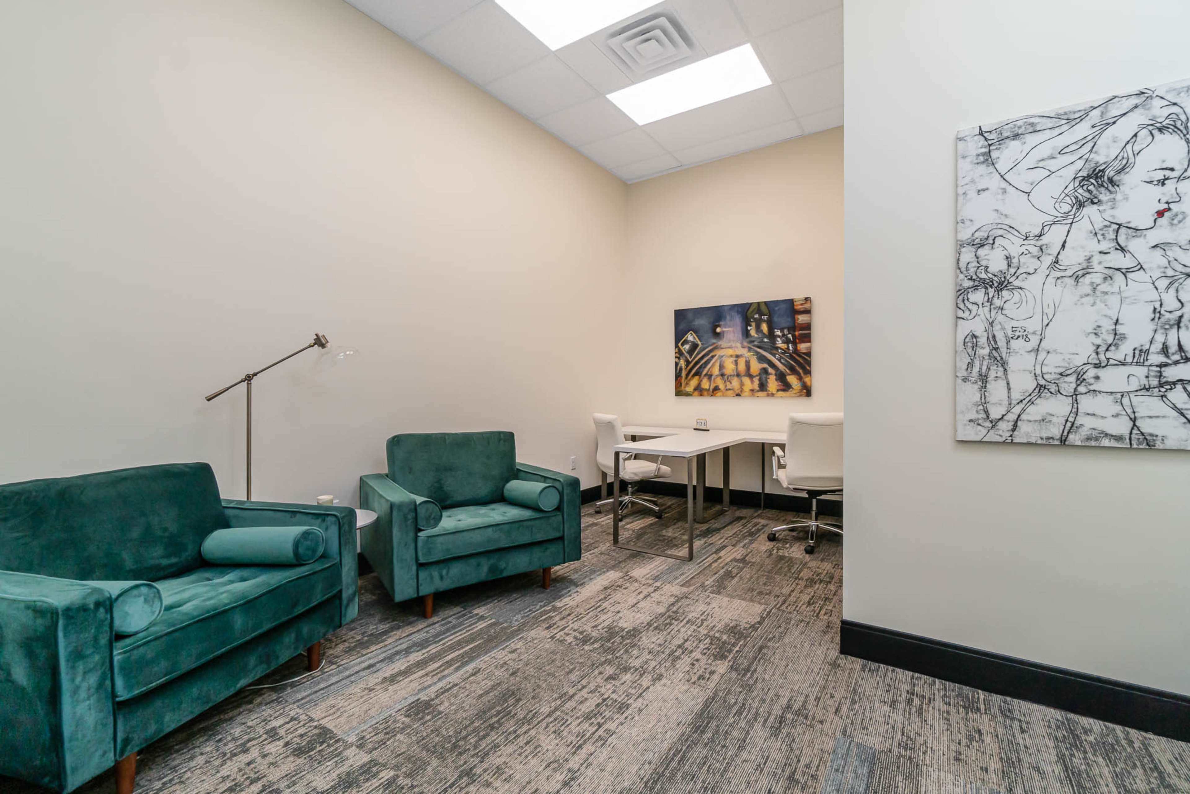 A small office space featuring two green armchairs, a modern desk with white chairs, and wall art on either side.