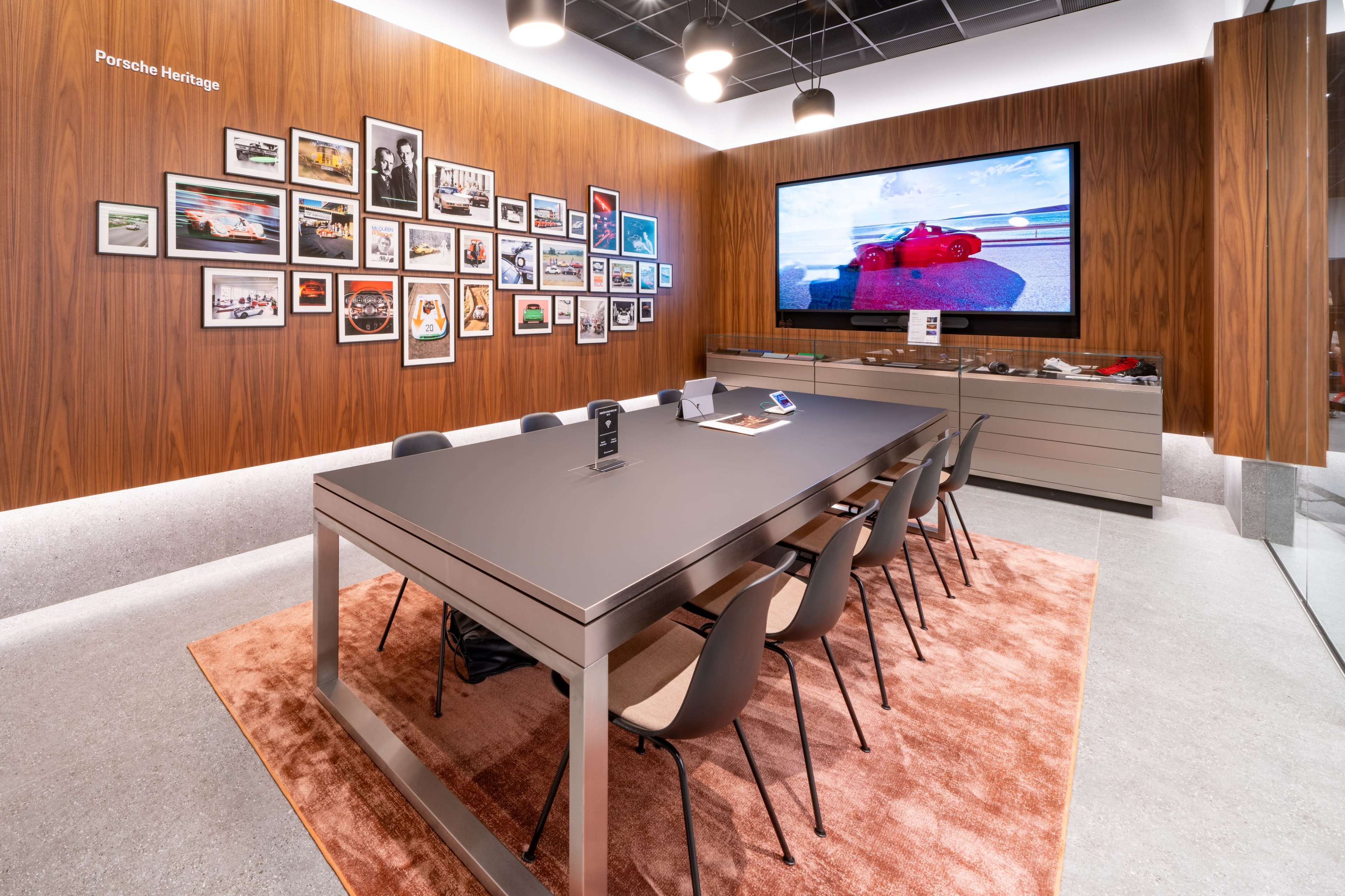 The image shows a modern conference room featuring a long table with chairs, a large television screen displaying images, and a wall adorned with framed photographs.