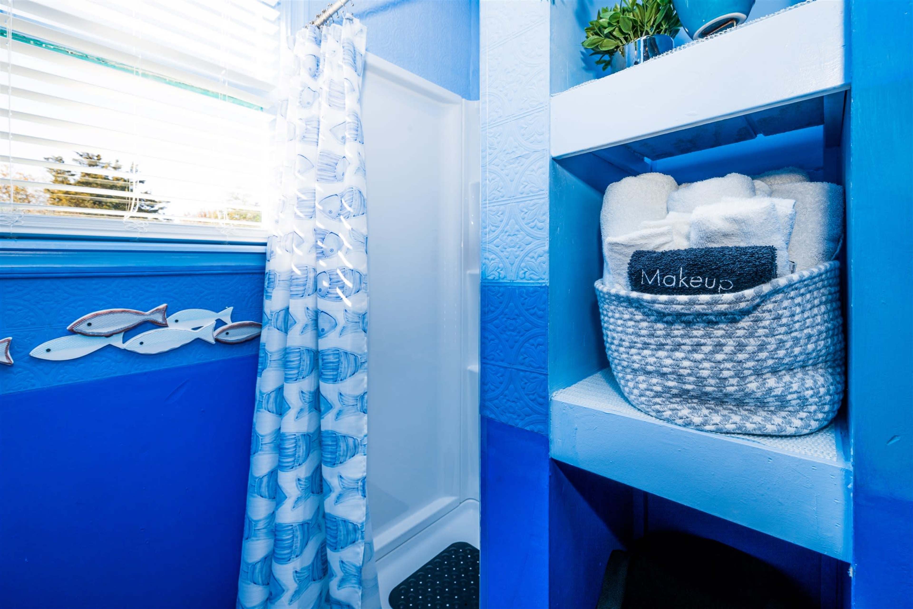A bright blue bathroom featuring a shower, a shelf with neatly folded towels, and a basket labeled "Makeup."