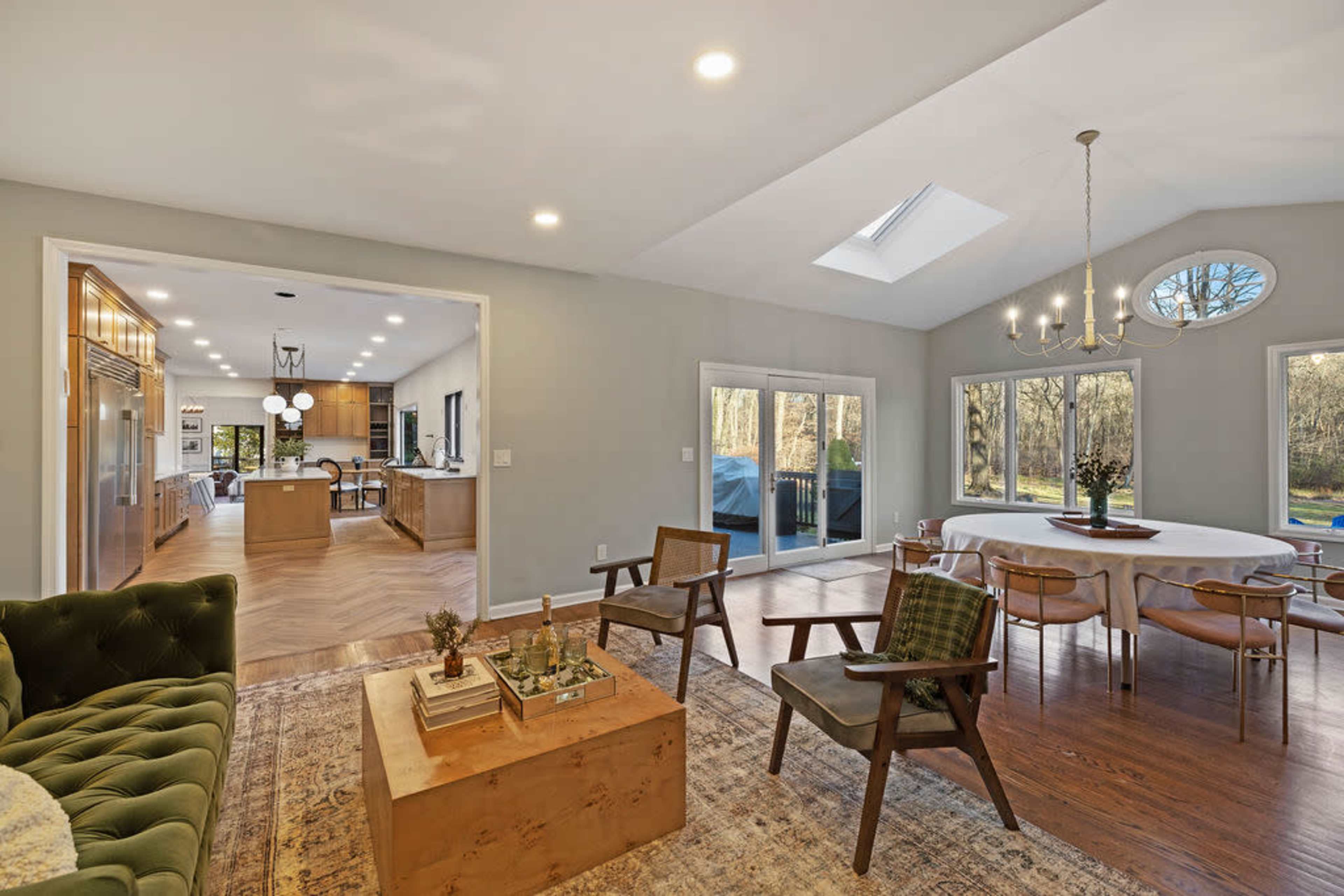 A spacious living area features a green velvet sofa, a wooden coffee table, and large windows allowing natural light, with a view of a dining area and kitchen.