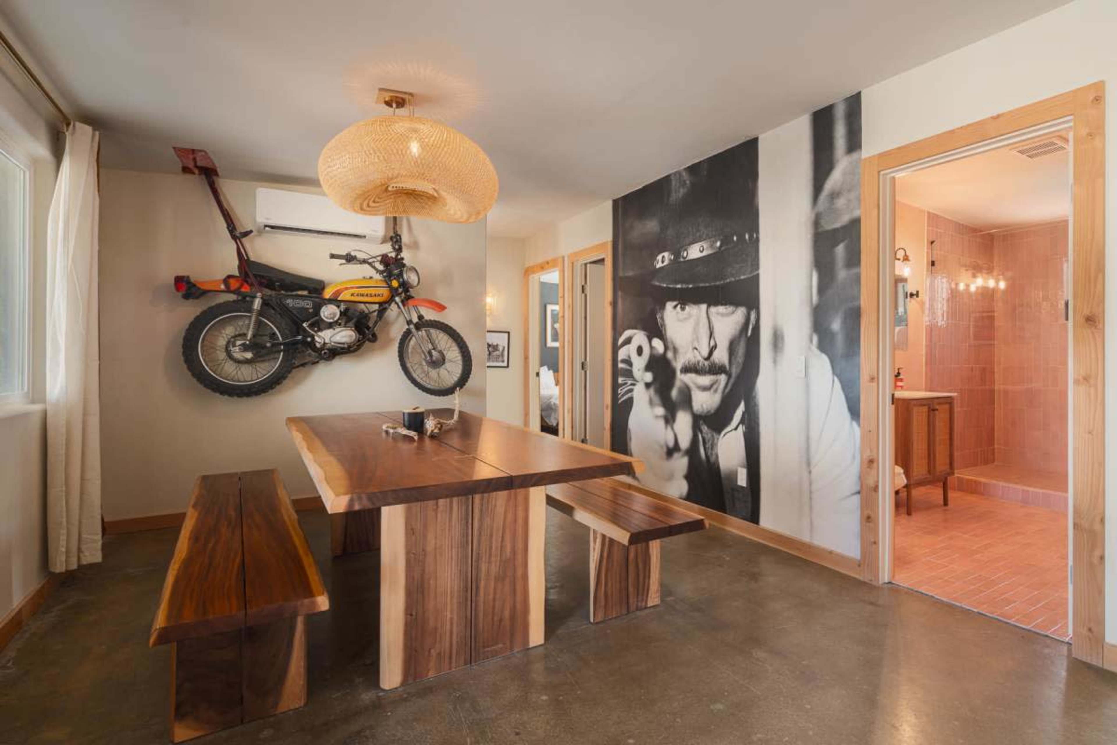 The image shows a dining area featuring a large wooden table, benches, a wall-mounted motorcycle, and a mural of a man pointing a gun.