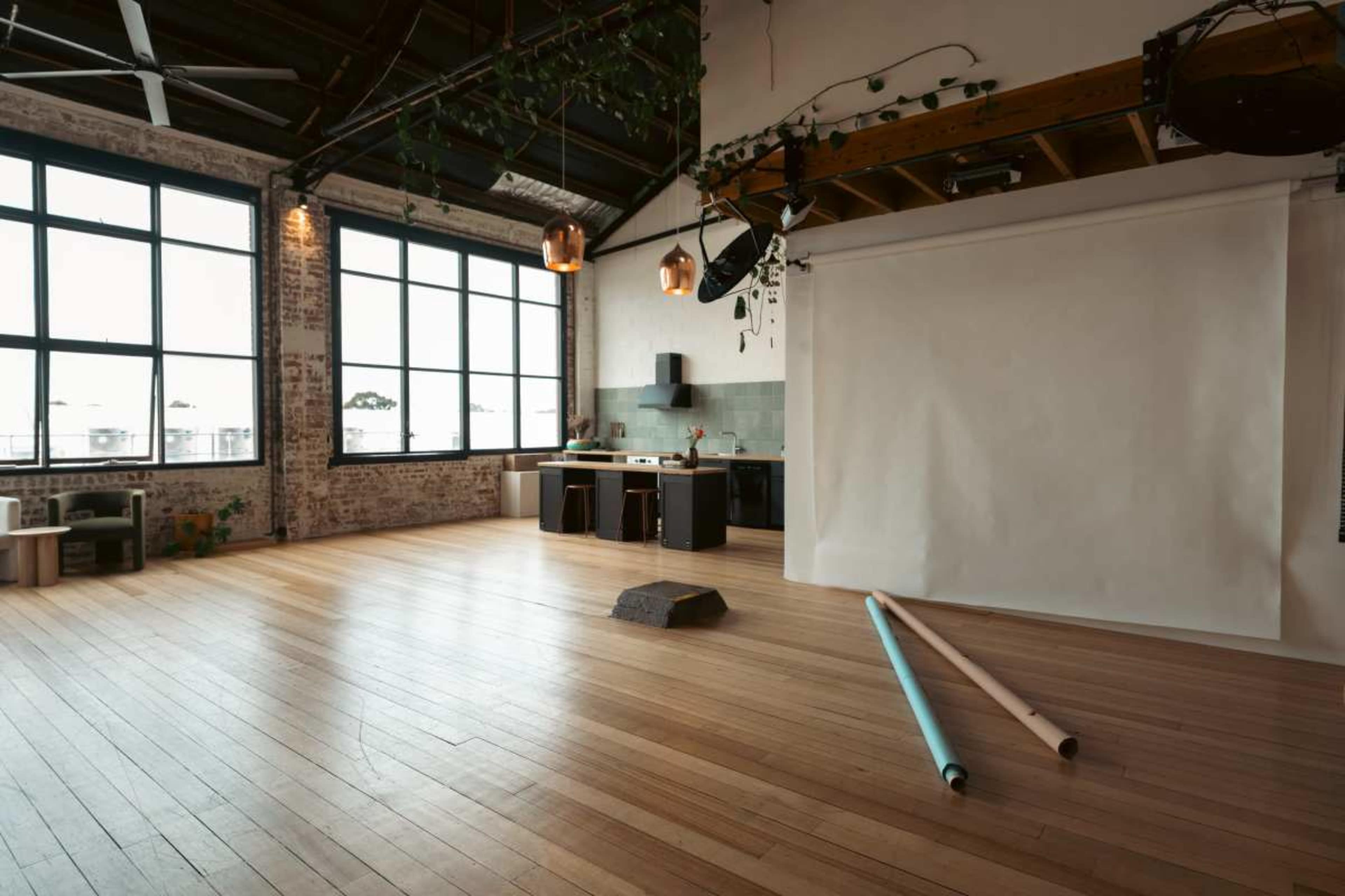 The image shows a spacious, well-lit studio with large windows, wooden flooring, a kitchen area, and a white backdrop with rolls of paper on the floor.