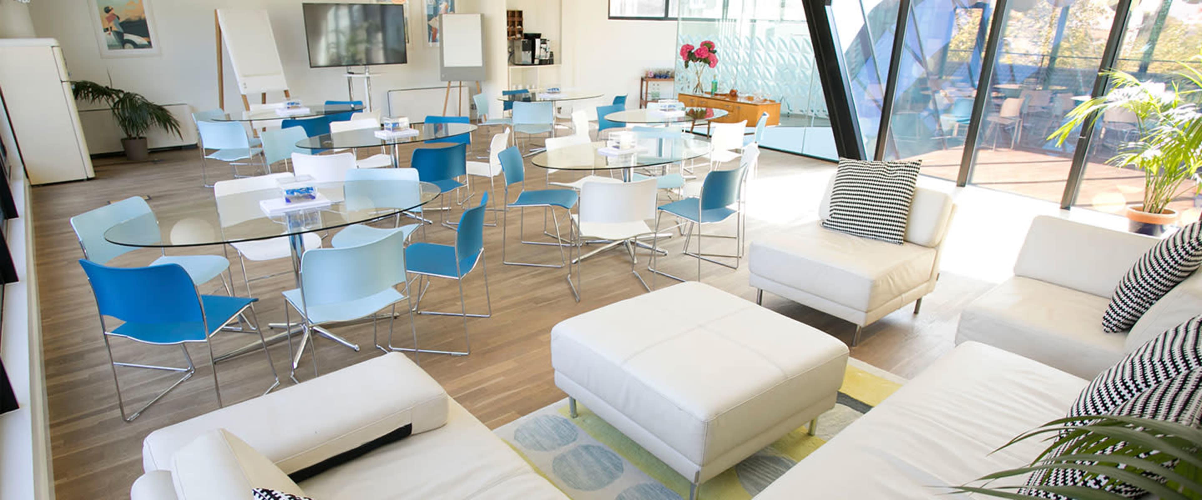 A modern meeting space equipped with glass tables, blue and white chairs, and a lounge area featuring white sofas.