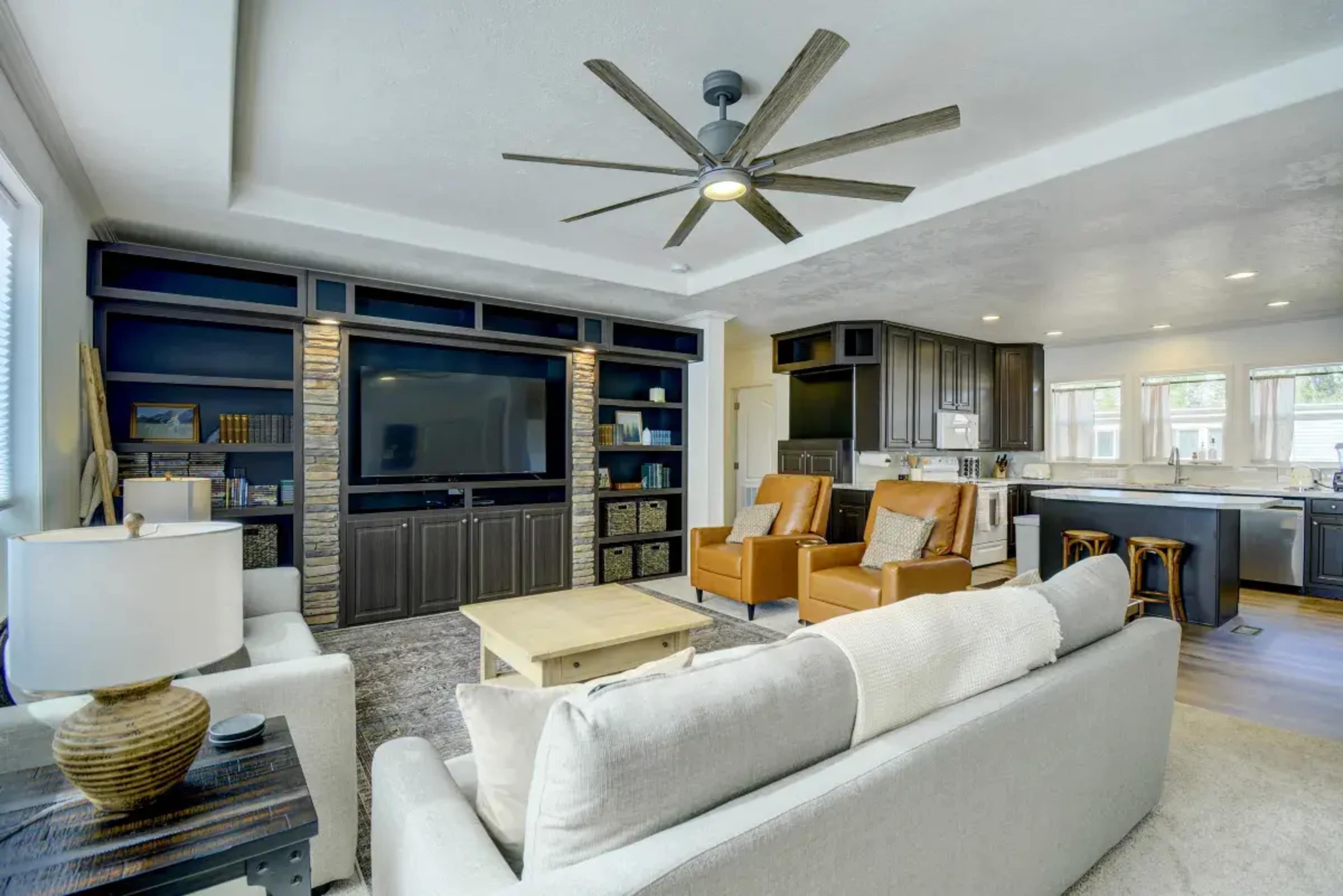 A modern living room with a large television on a built-in shelf, two leather recliners, and a light-colored sofa, all arranged around a central coffee table.