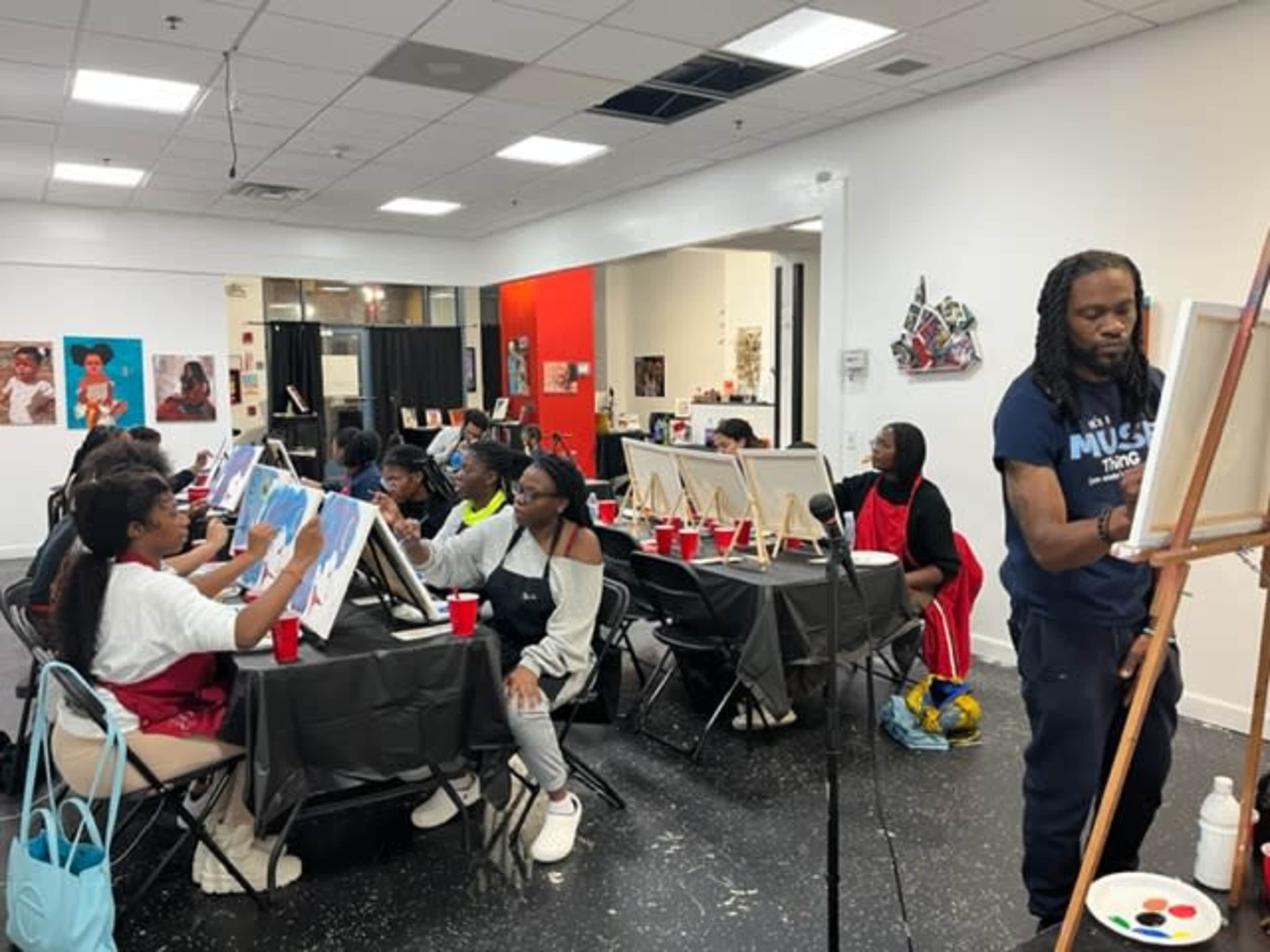 A group of individuals is attending a painting class, with an instructor demonstrating on a canvas in the front while participants work on their own artworks at easels.