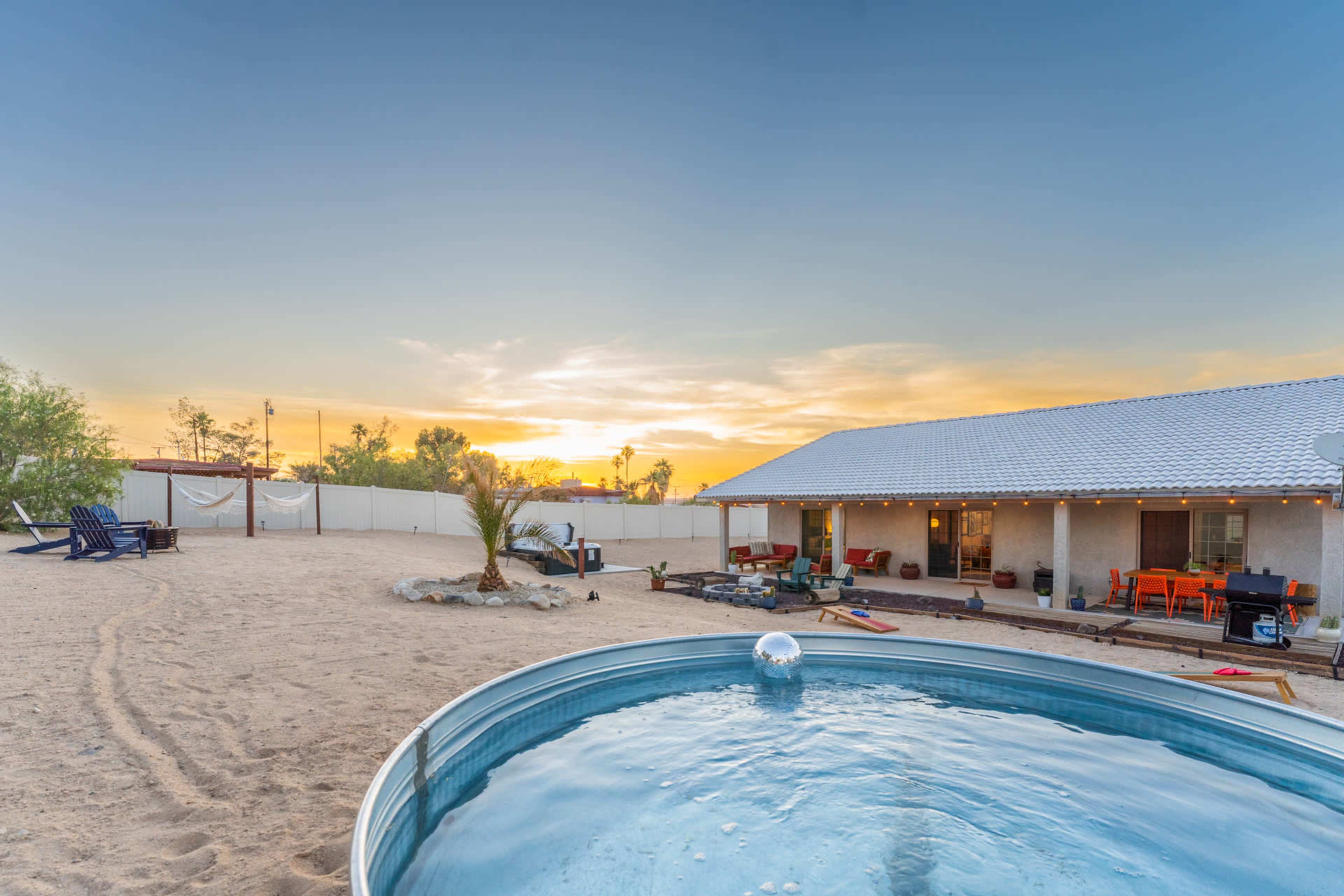 The image shows a backyard with a round pool, sandy ground, and a house with a patio overlooking a colorful sunset.