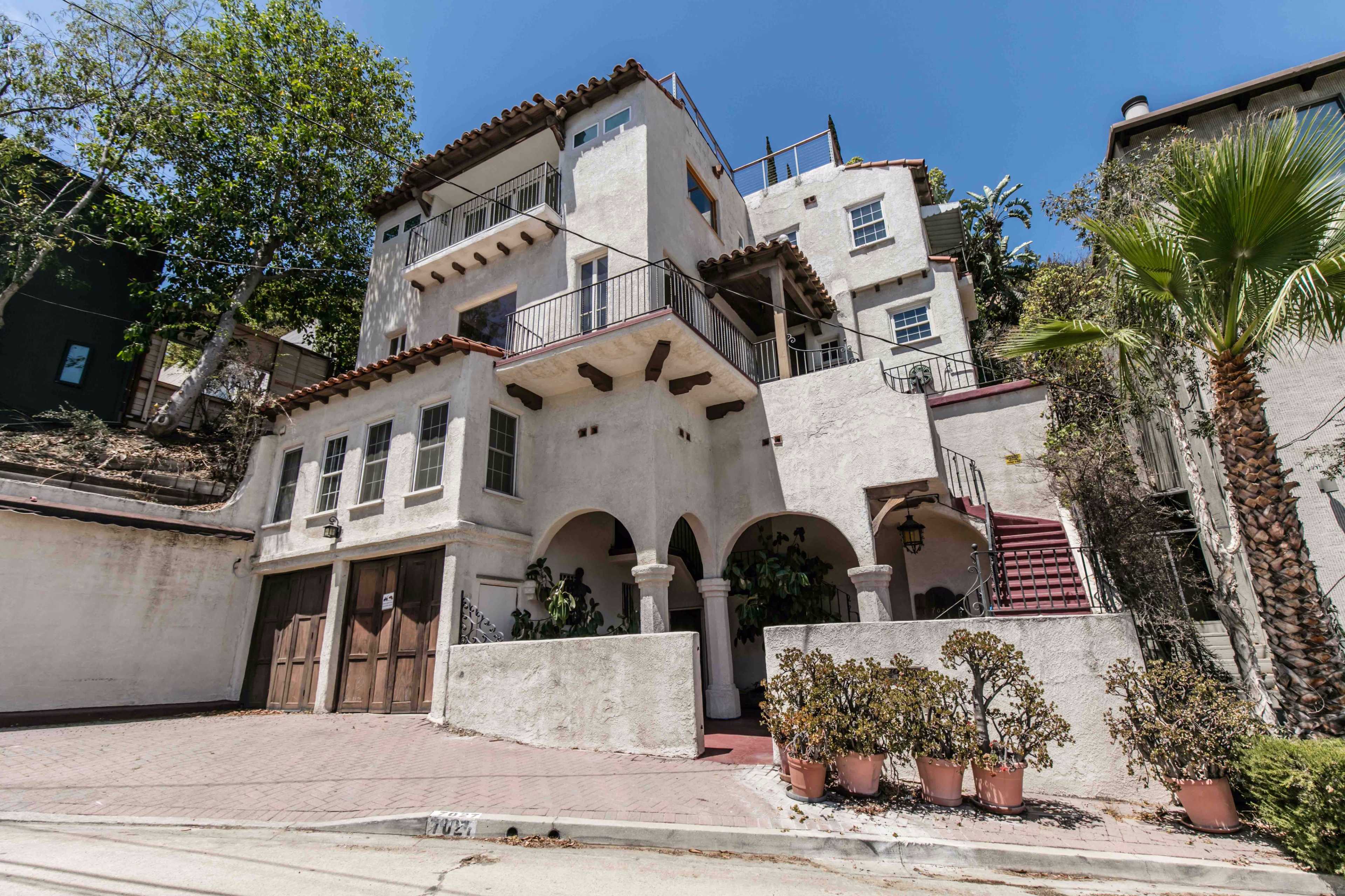 The image shows a three-story stucco house with arched doorways, a red-tiled roof, and a driveway lined with potted plants.