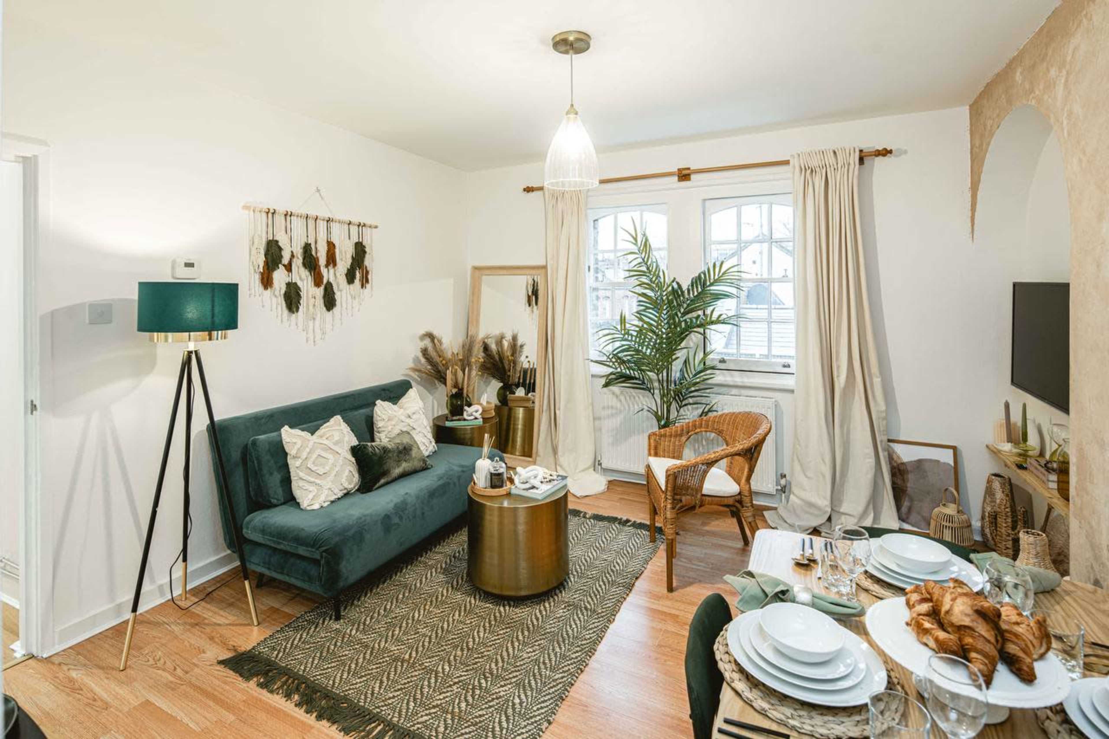 The image shows a cozy living area featuring a green velvet sofa, a round gold coffee table, and a woven chair, all set against a light-colored wall with decorative elements.