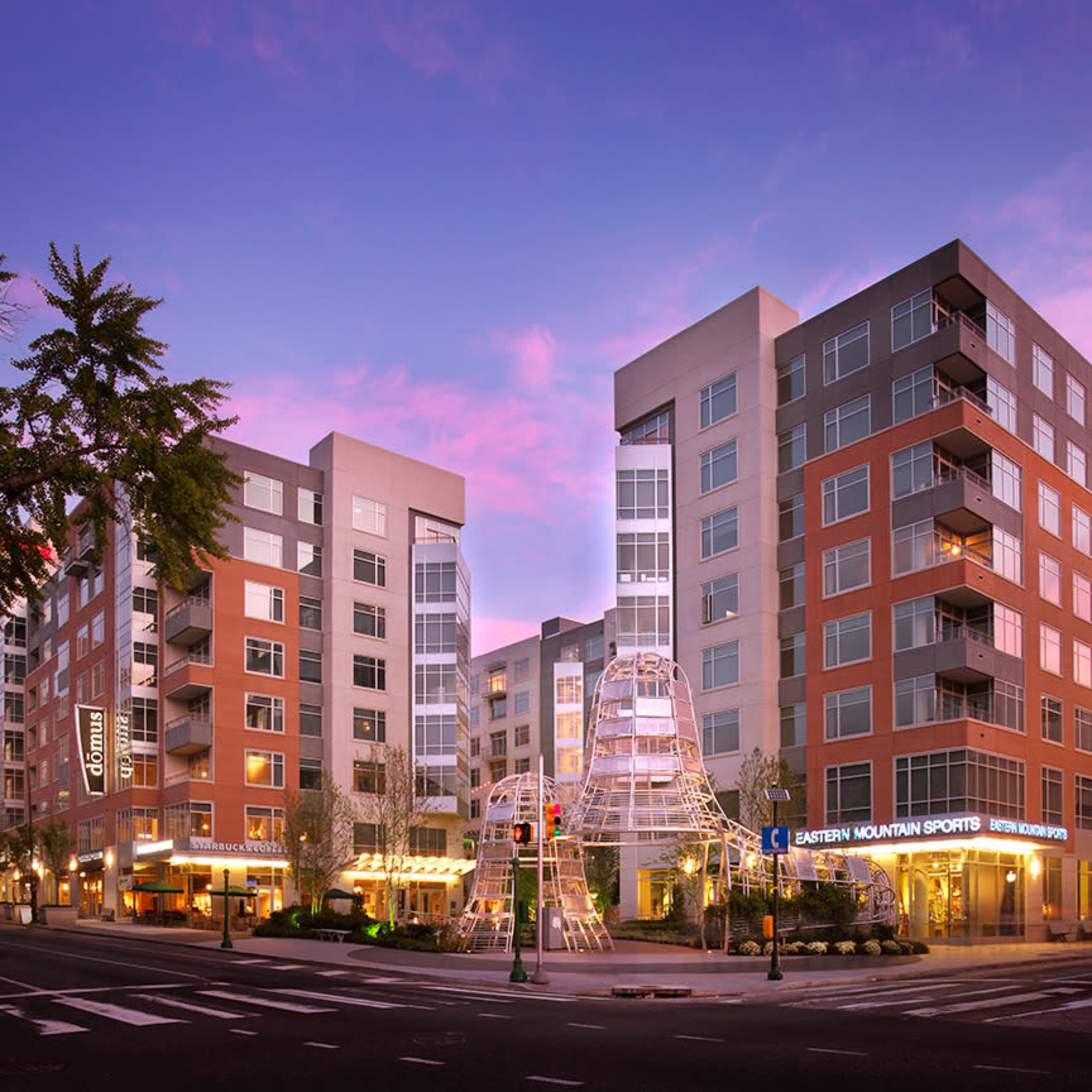 The image shows a modern apartment complex with multiple buildings and a children's play structure at the intersection of two streets during sunset.