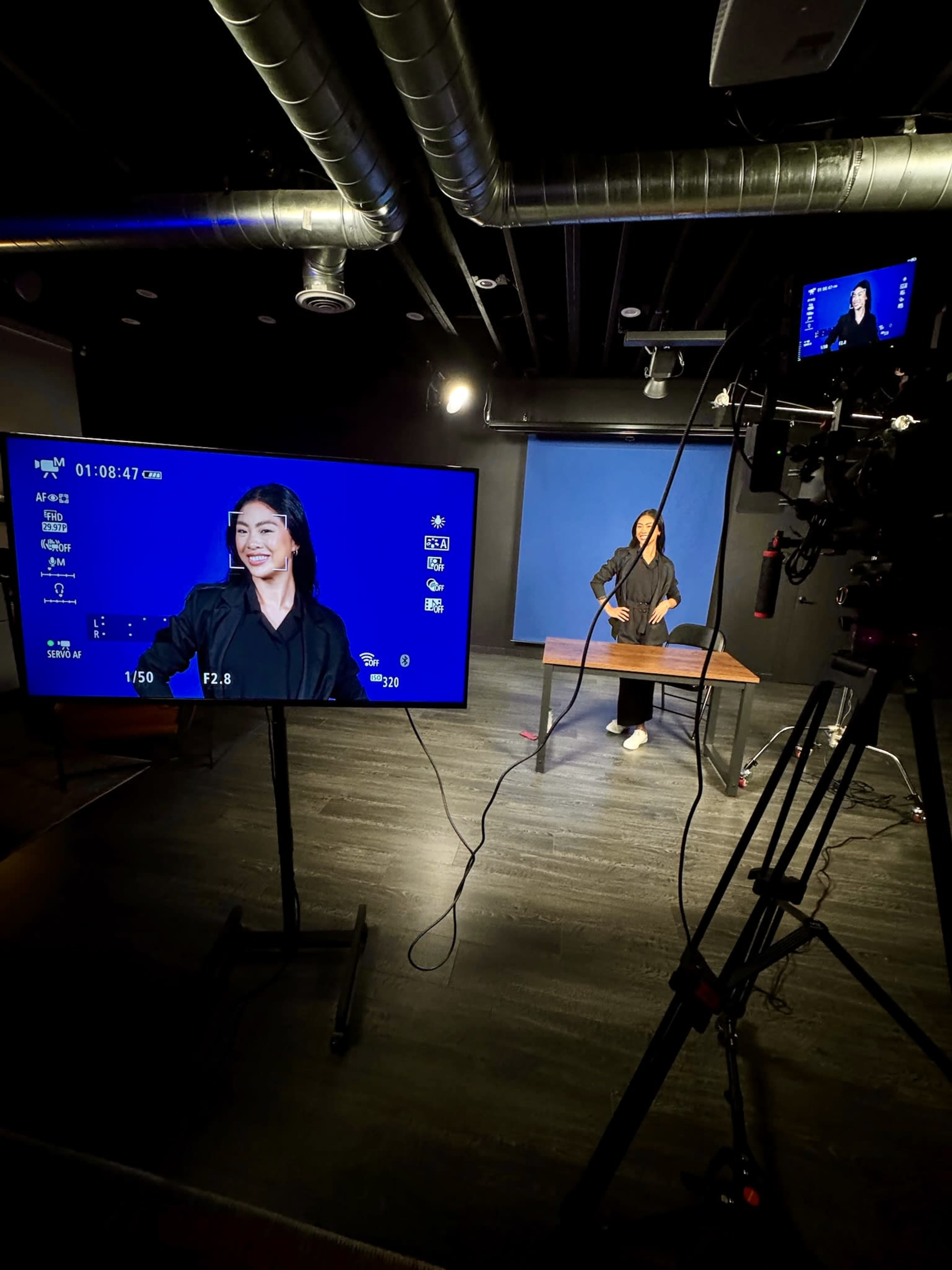 A person in a black outfit stands at a table while being recorded in a studio with professional lighting and camera equipment.