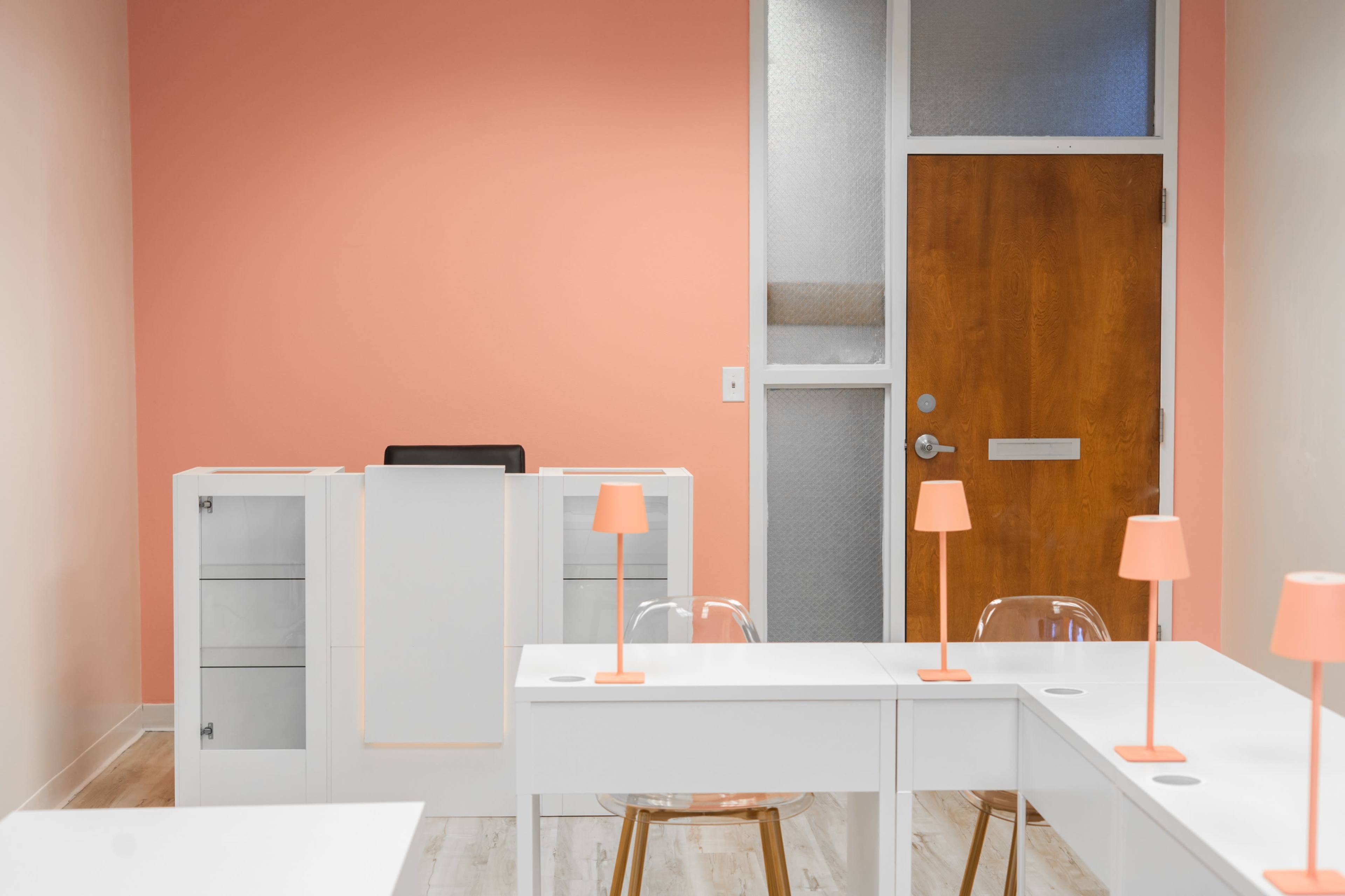 The image shows a minimalist office space with white desks and orange lamps against a light peach-colored wall and a wooden door.