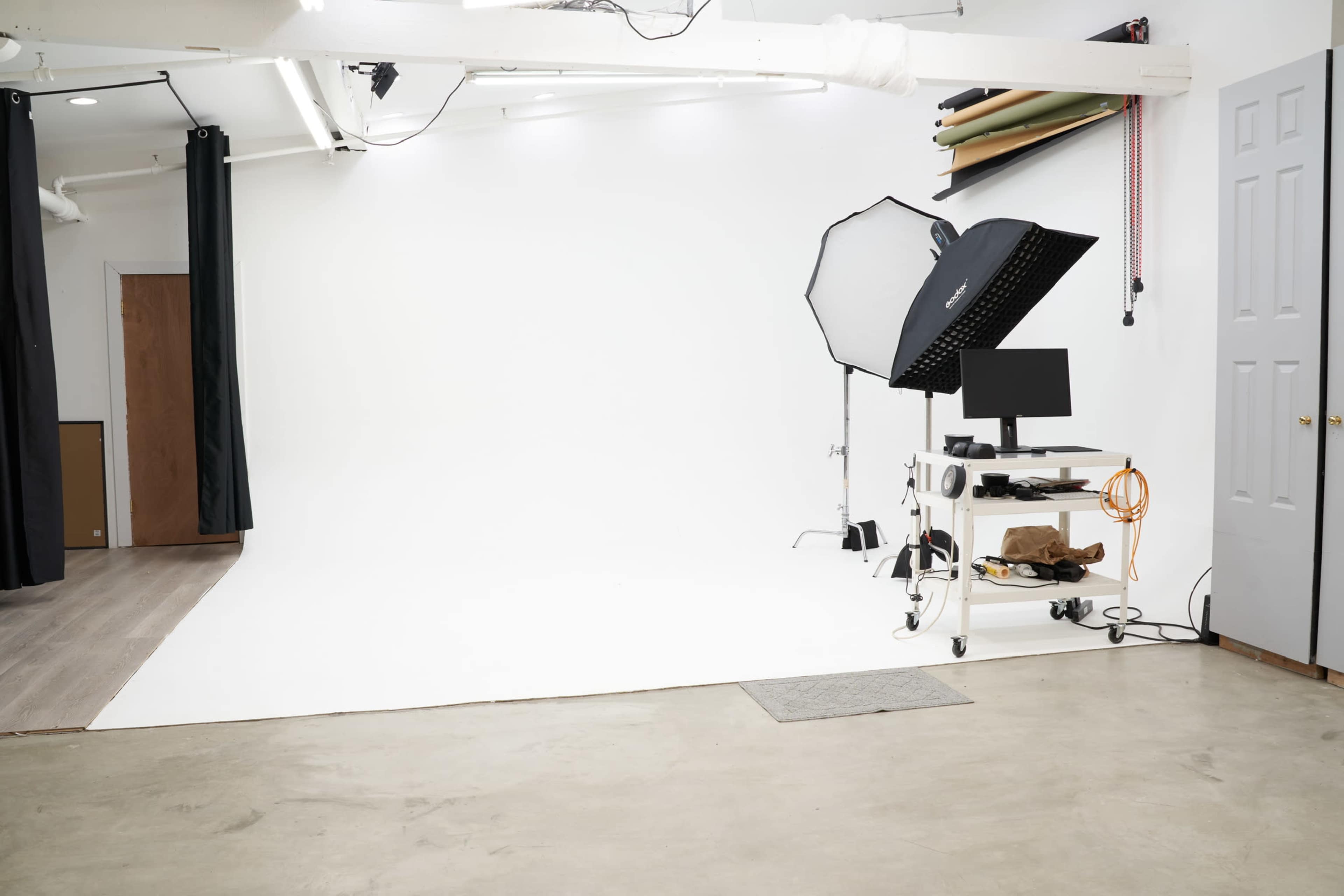 The image shows a blank photography studio with a white backdrop, lighting equipment, and a computer on a mobile cart.
