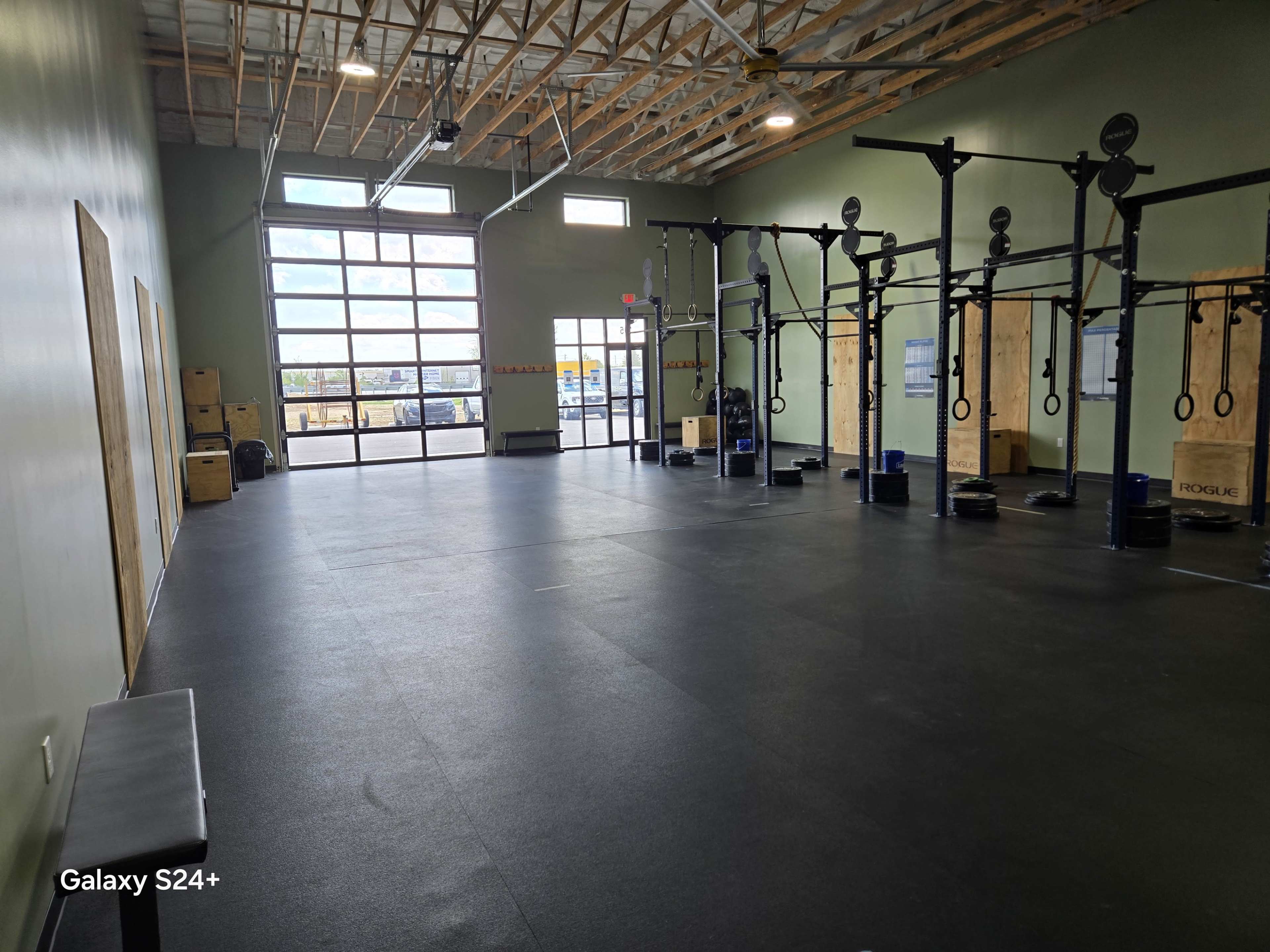 The image shows a spacious gym interior with exercise equipment, including pull-up bars and wooden boxes, alongside large windows letting in natural light.