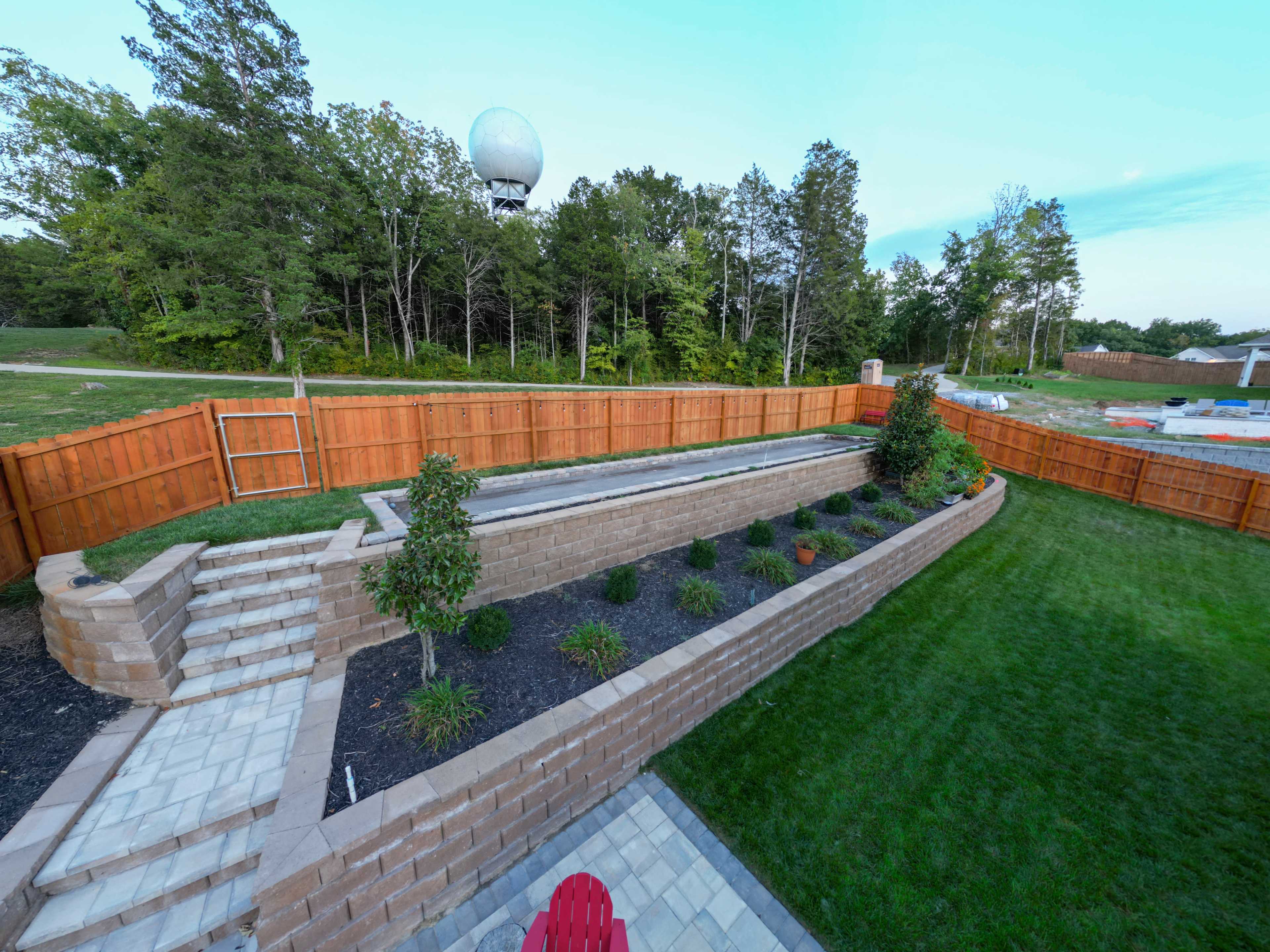 The image shows a landscaped backyard with a stone staircase leading to a raised garden bed bordered by wooden fencing and trees in the background.