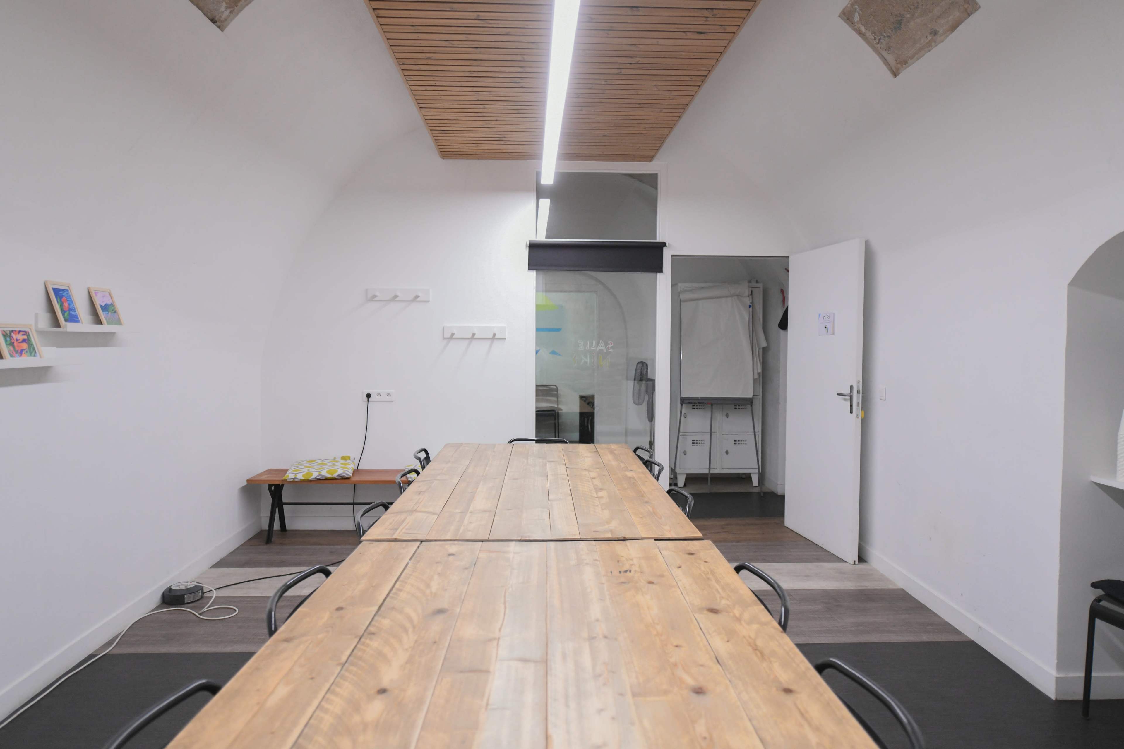 A long wooden table with black chairs is centered in a well-lit room featuring a simple design and an adjoining door to another space.
