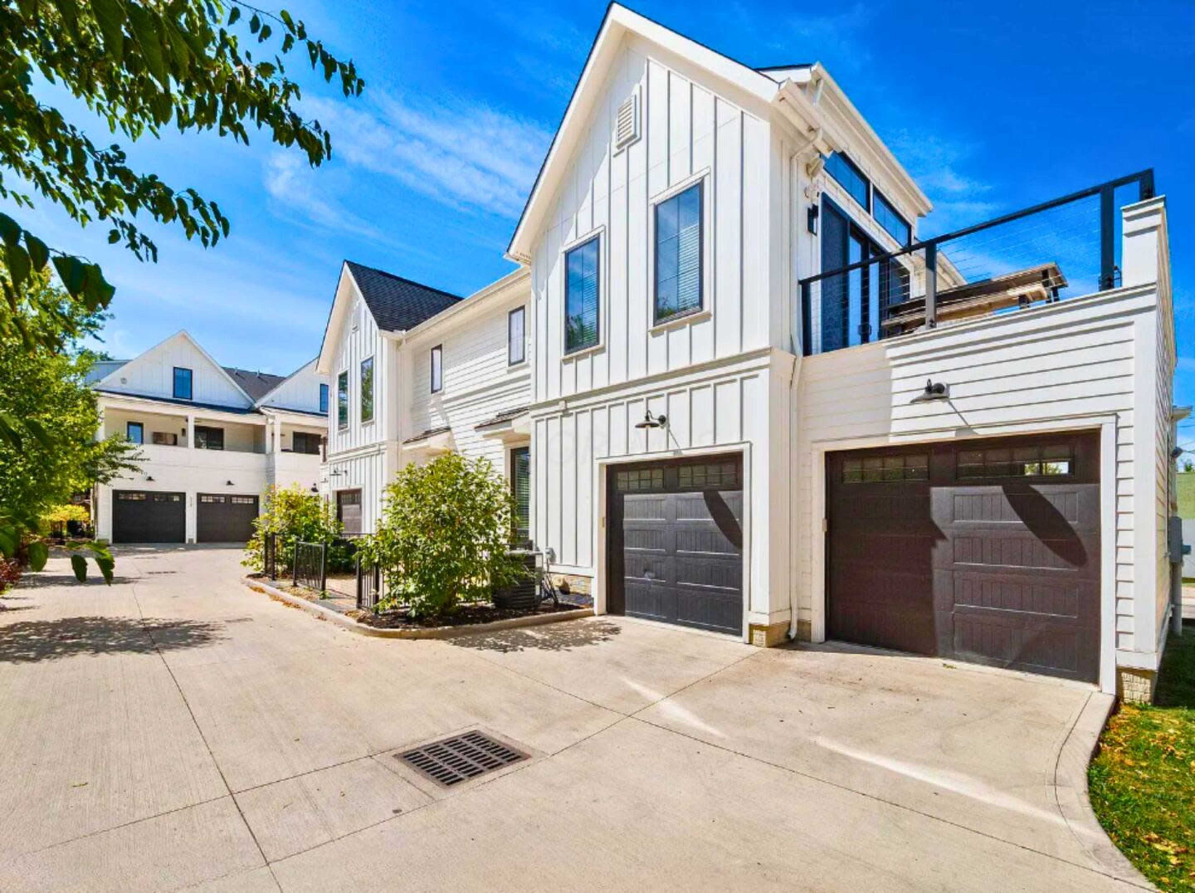 The image shows a modern white house with black garage doors, situated in a residential area with clear blue skies and greenery.