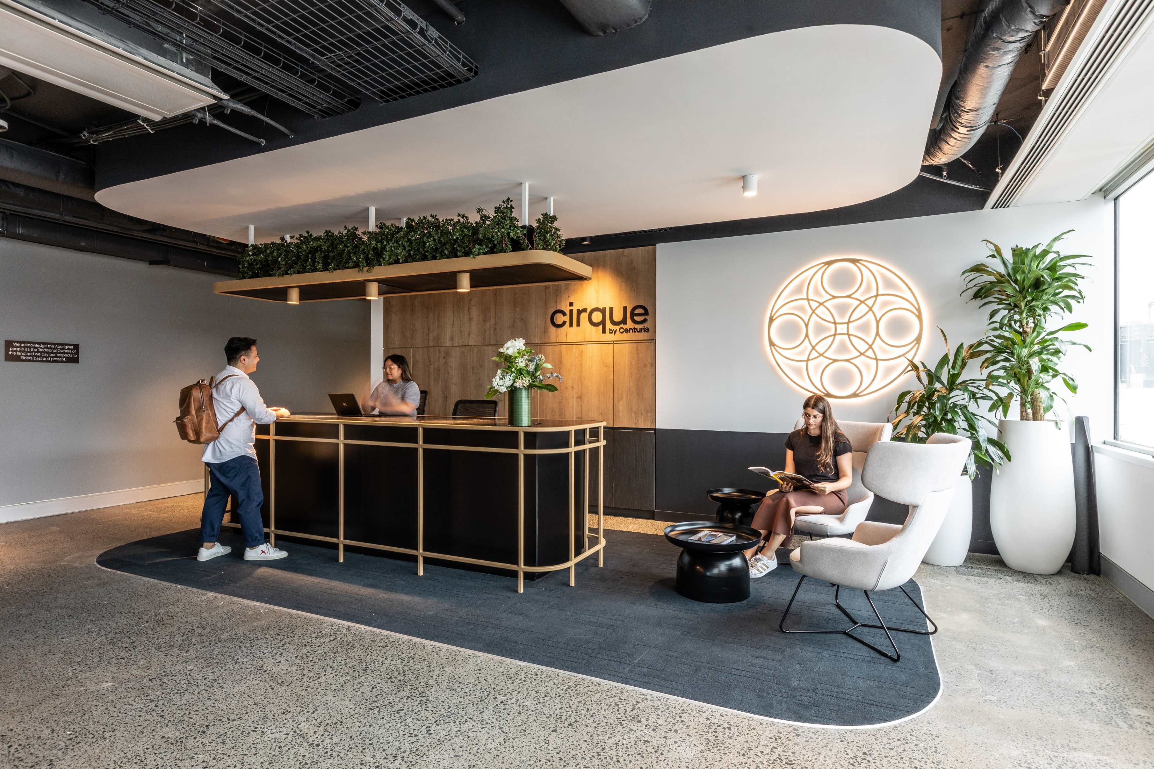 The image shows a contemporary reception area with a wooden desk, a potted plant, and a woman sitting in a chair, reading.