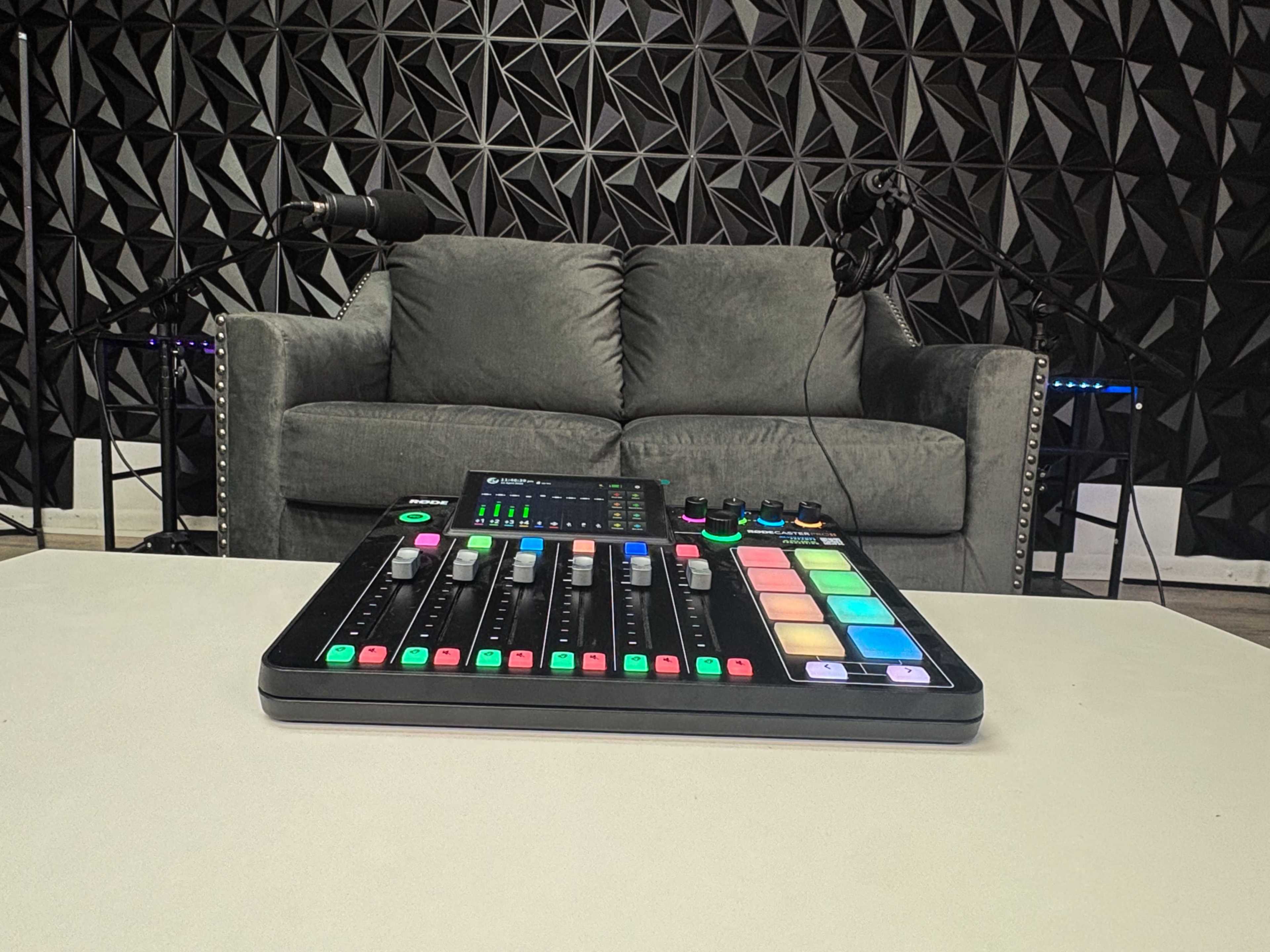 The image shows a sound mixing board on a table in front of a gray couch with two microphones positioned on either side in a studio setting.