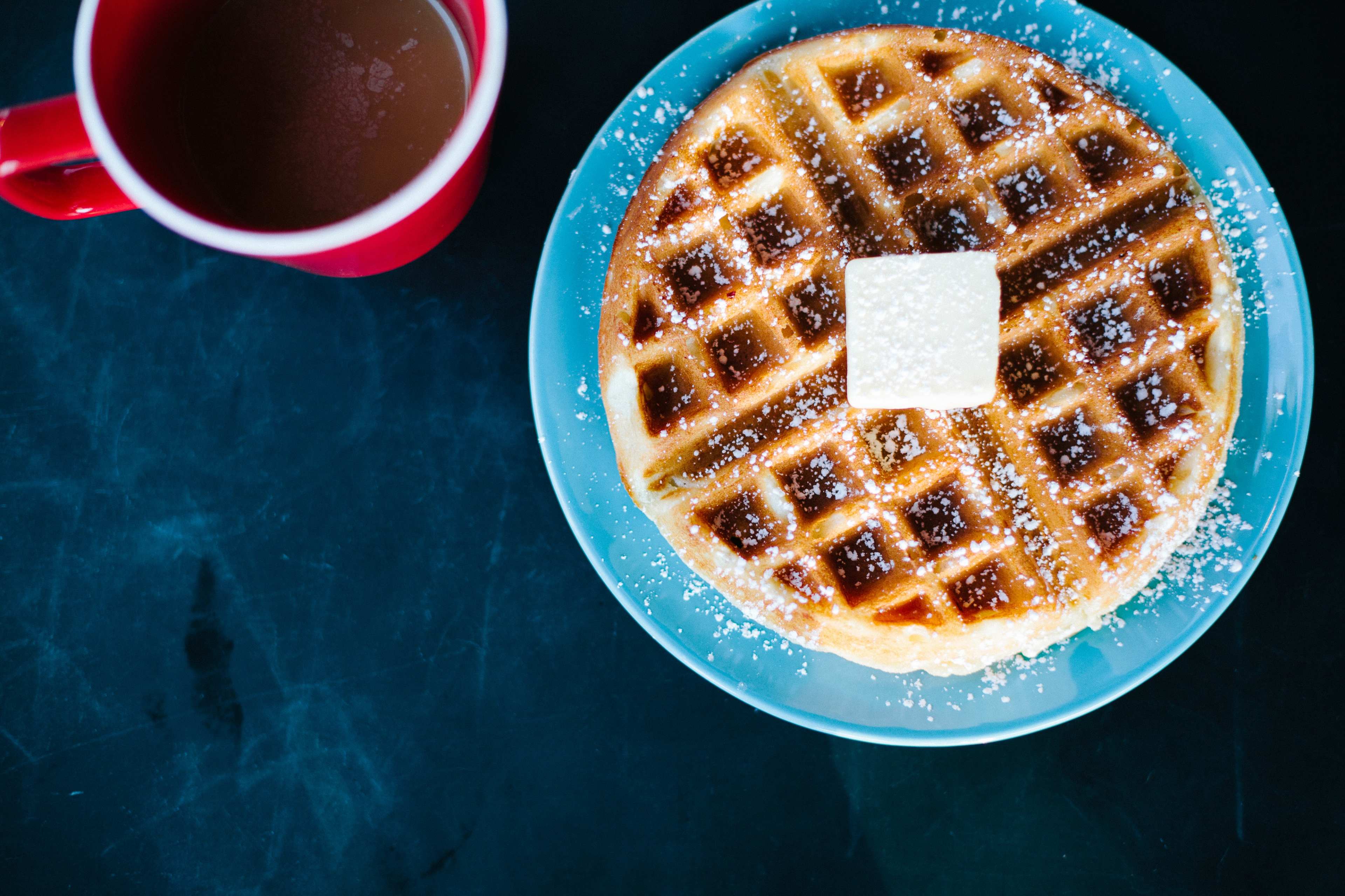 A golden waffle topped with a pat of butter sits on a blue plate next to a red cup filled with coffee.