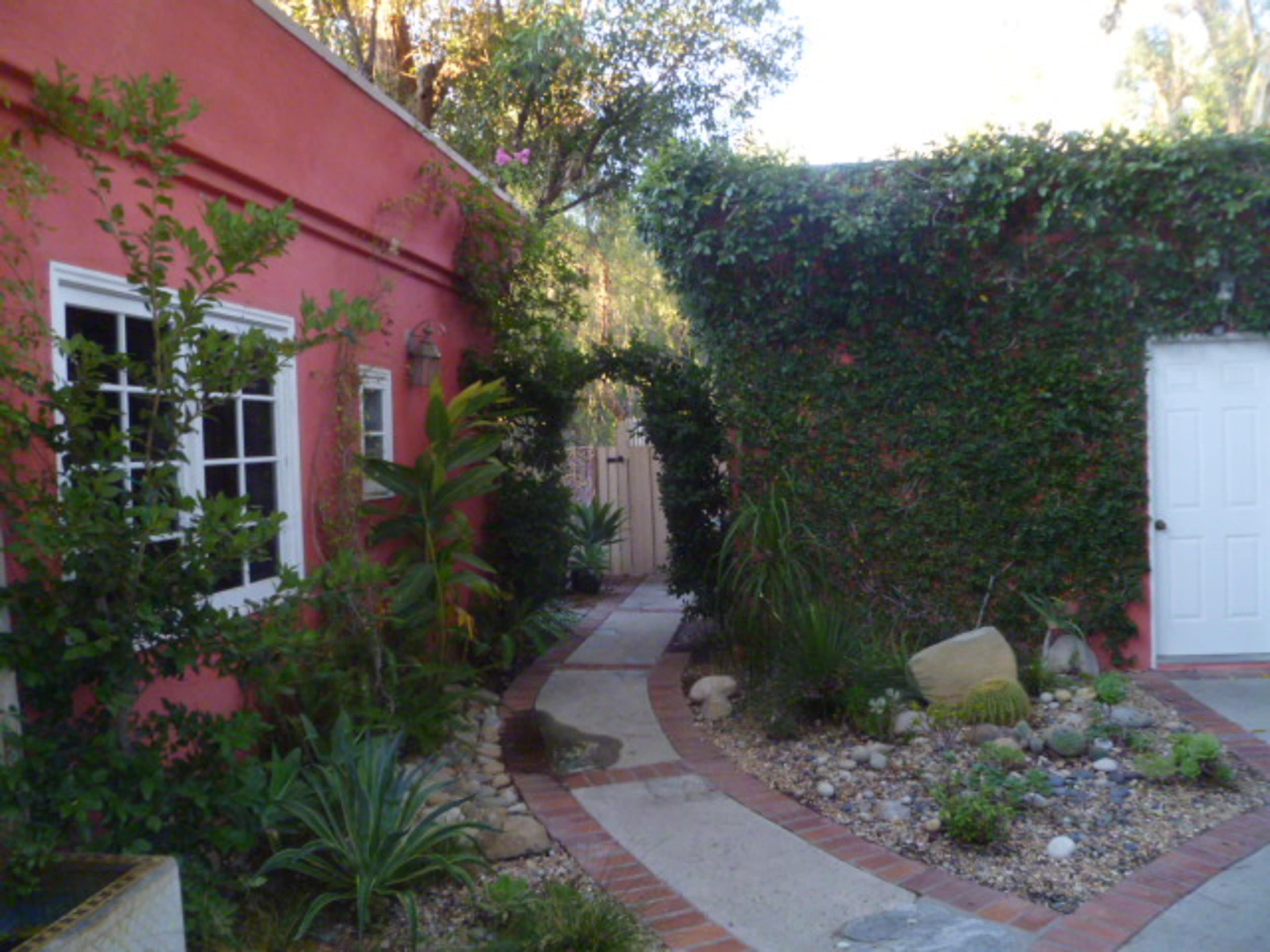 A winding stone path leads through a garden with lush greenery and a pink building on one side.