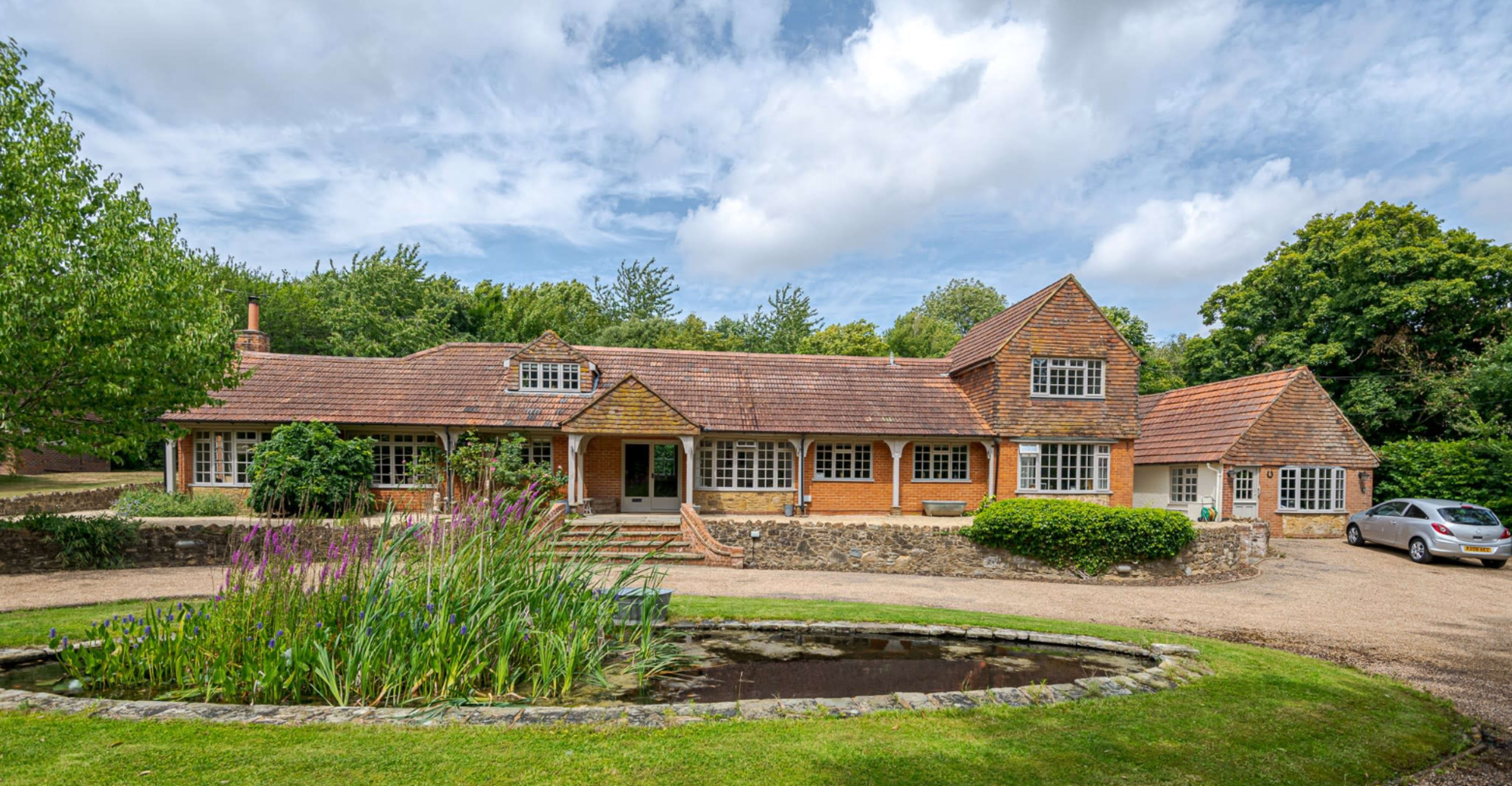 The image shows a large brick house with a tiled roof, surrounded by well-maintained gardens and a pond in the foreground.