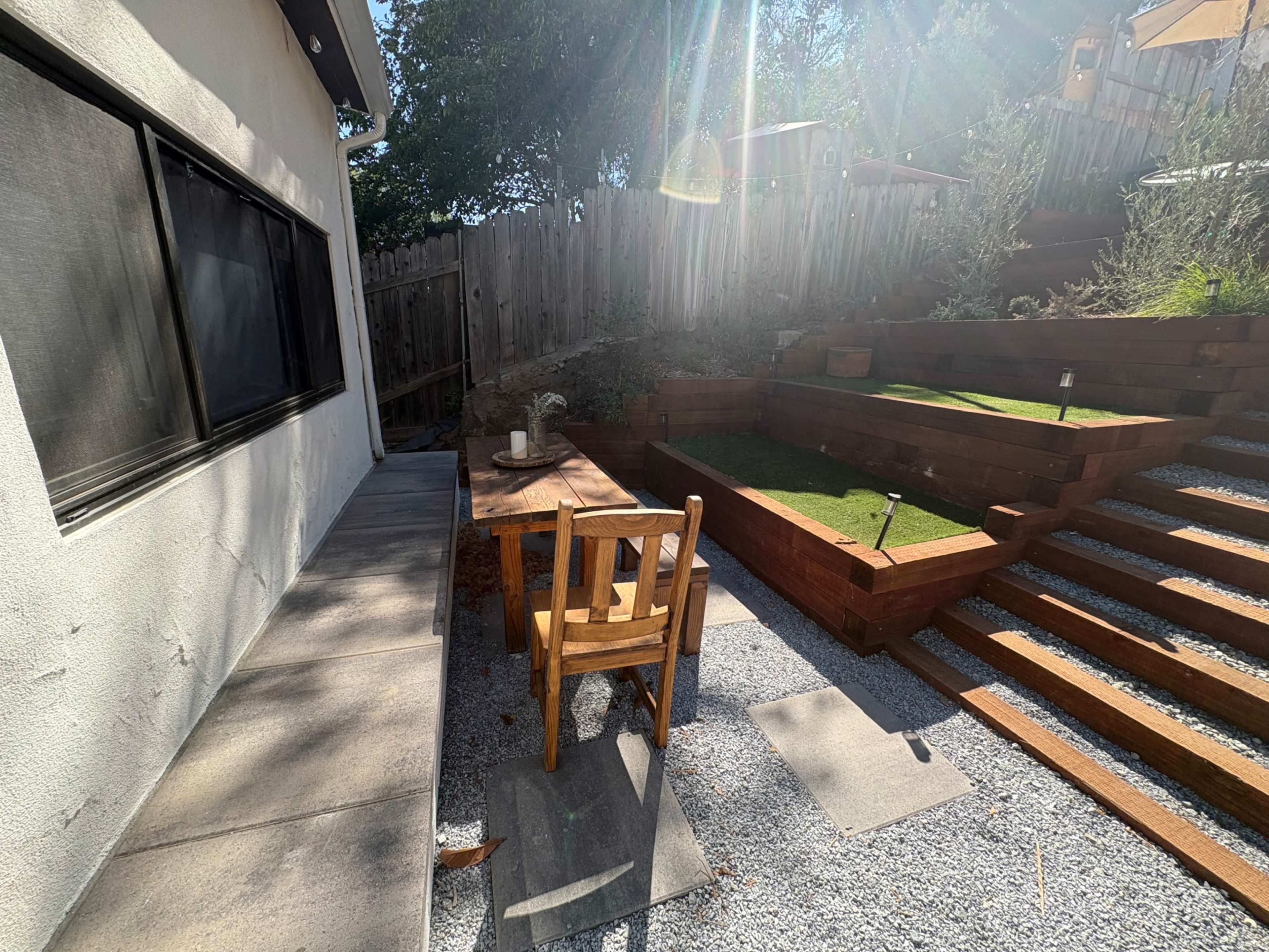 A wooden table and chair are positioned next to a gravel pathway, leading to a terraced garden with wooden steps and a small green lawn.