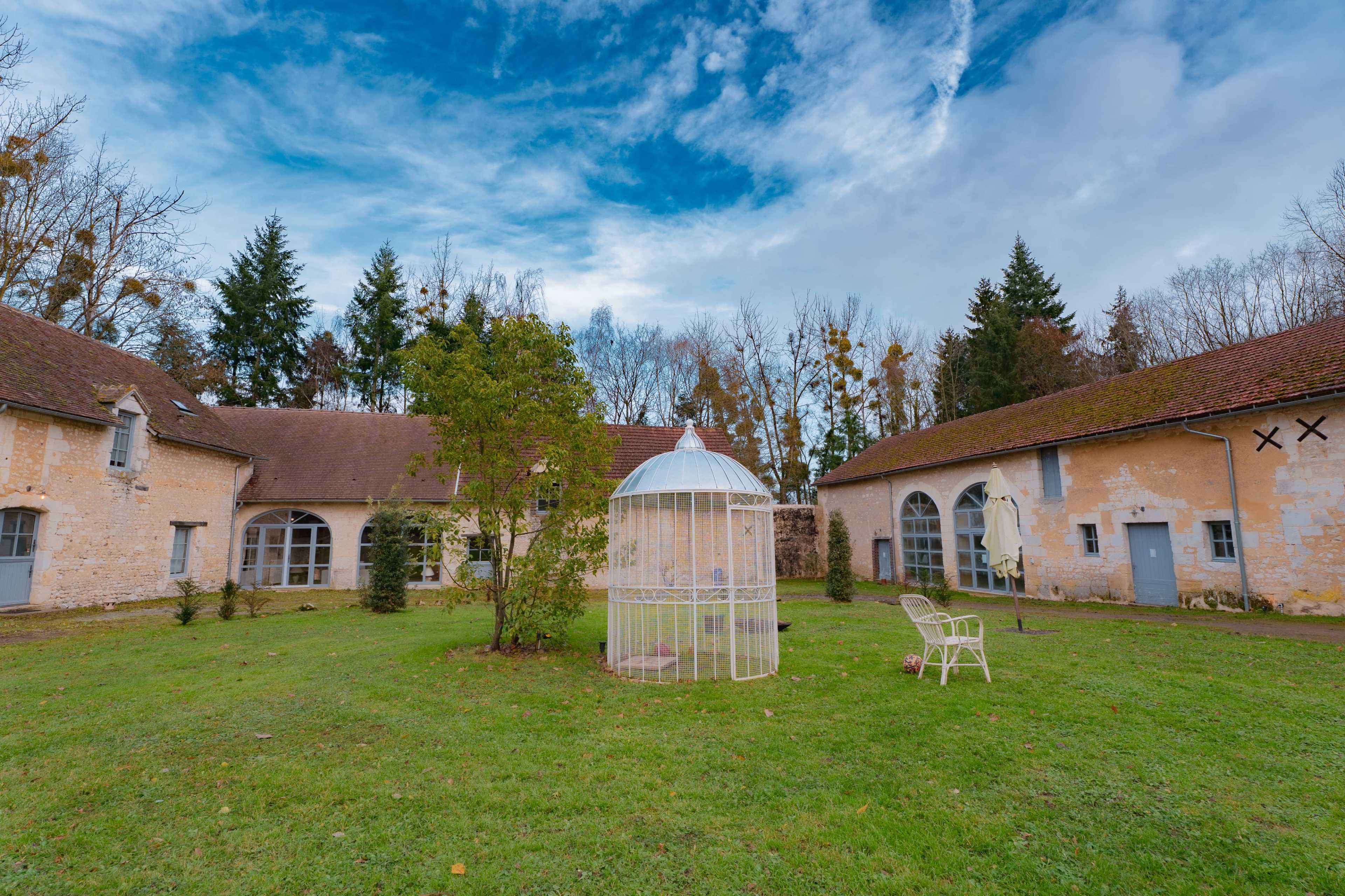 The image shows a white birdcage standing on a grassy area surrounded by buildings with stone walls and a forest backdrop under a cloudy sky.