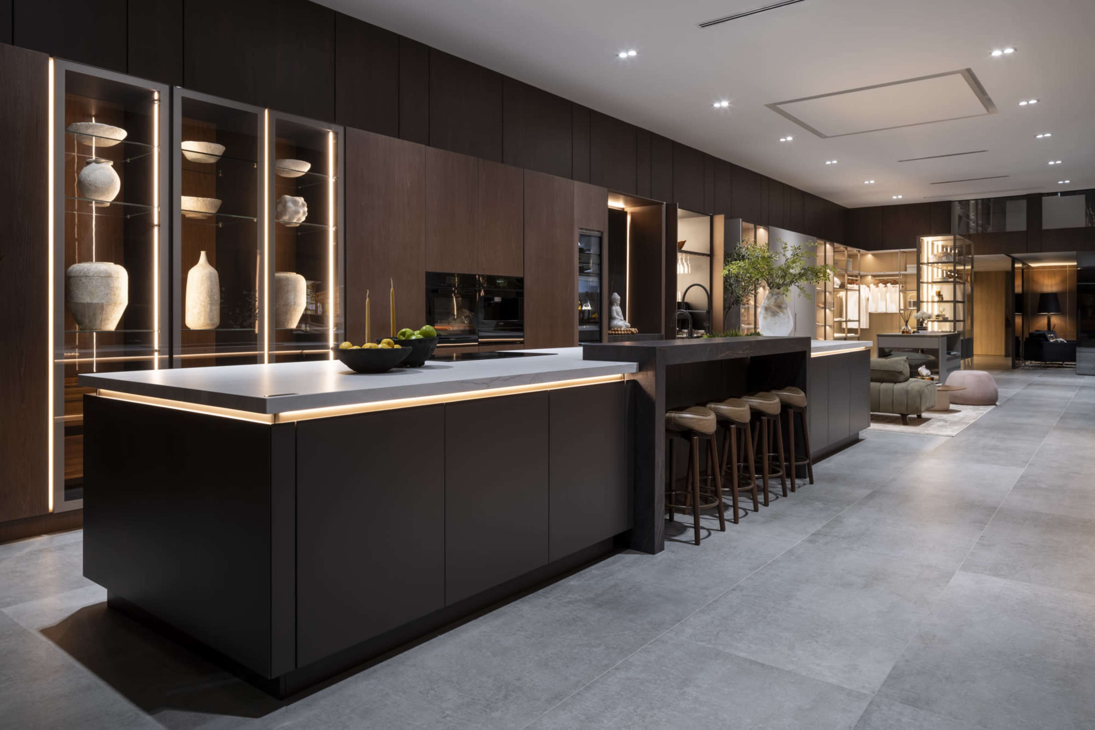 A modern kitchen features dark cabinetry, a large central island with seating, and illuminated glass displays showcasing decorative items.