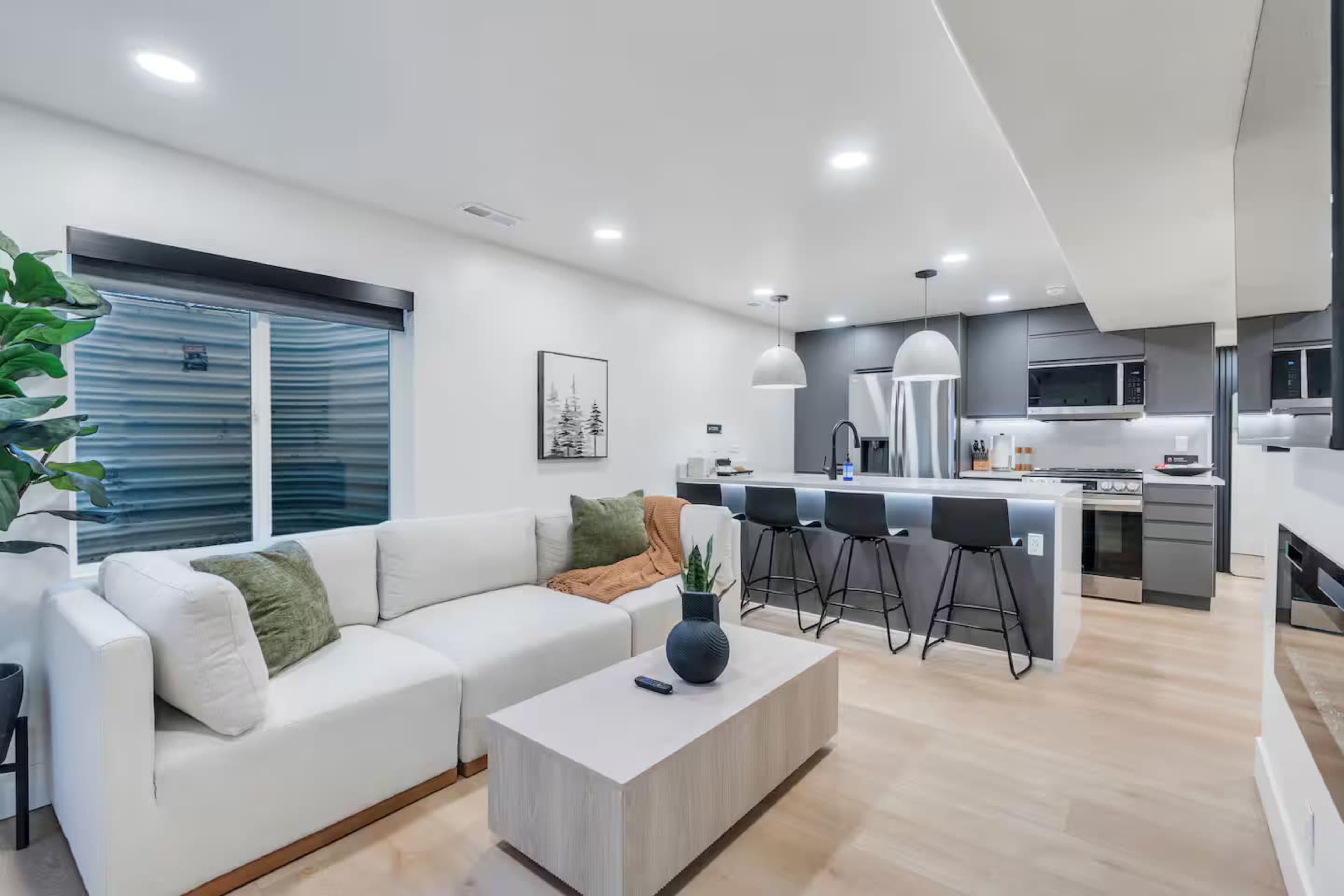 The image shows a modern living space featuring a white sectional sofa, a coffee table, and a kitchen area with bar stools and stainless steel appliances.