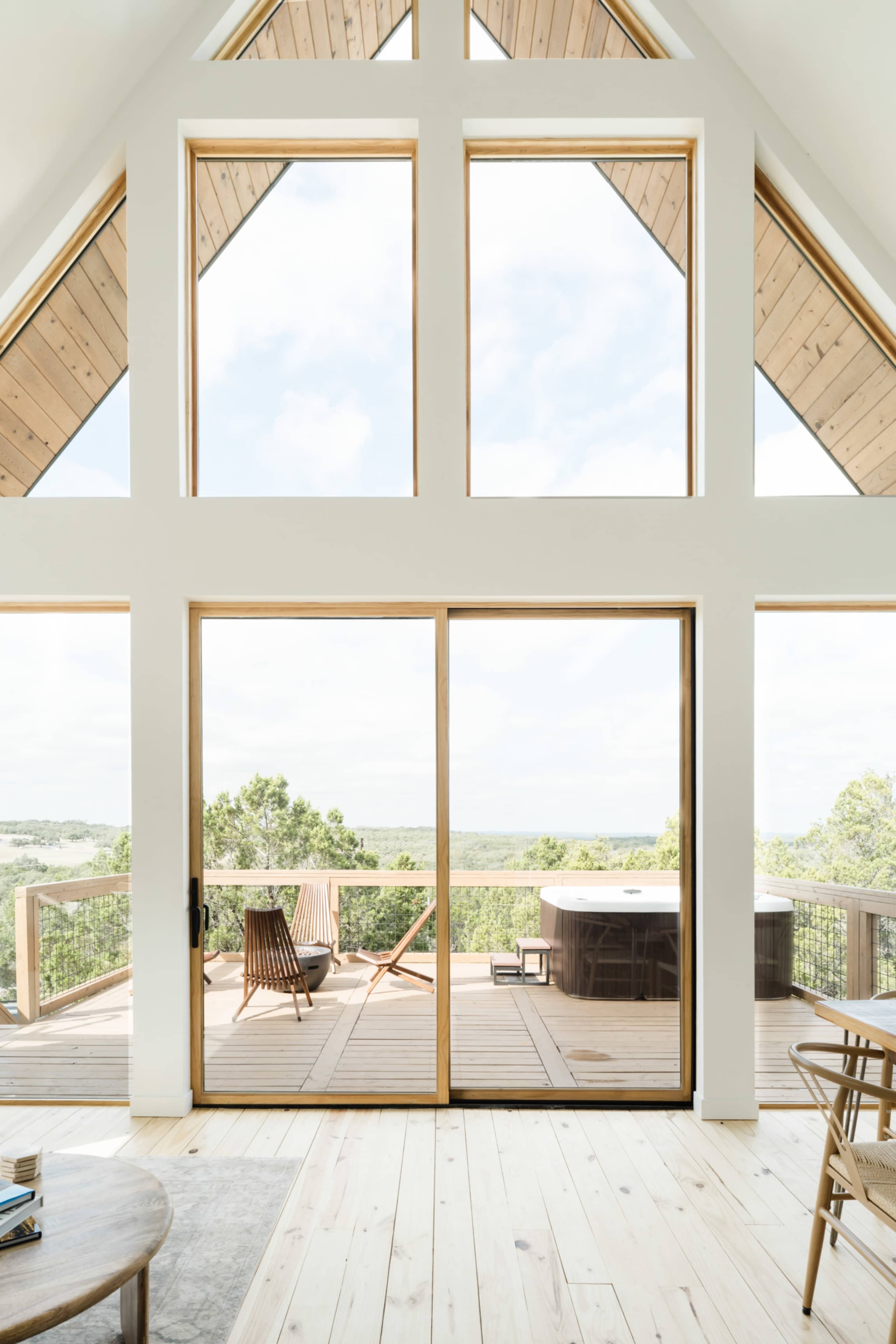 A spacious room with large windows overlooking a wooden deck that includes a hot tub and chairs, situated in a house with a high A-frame ceiling.