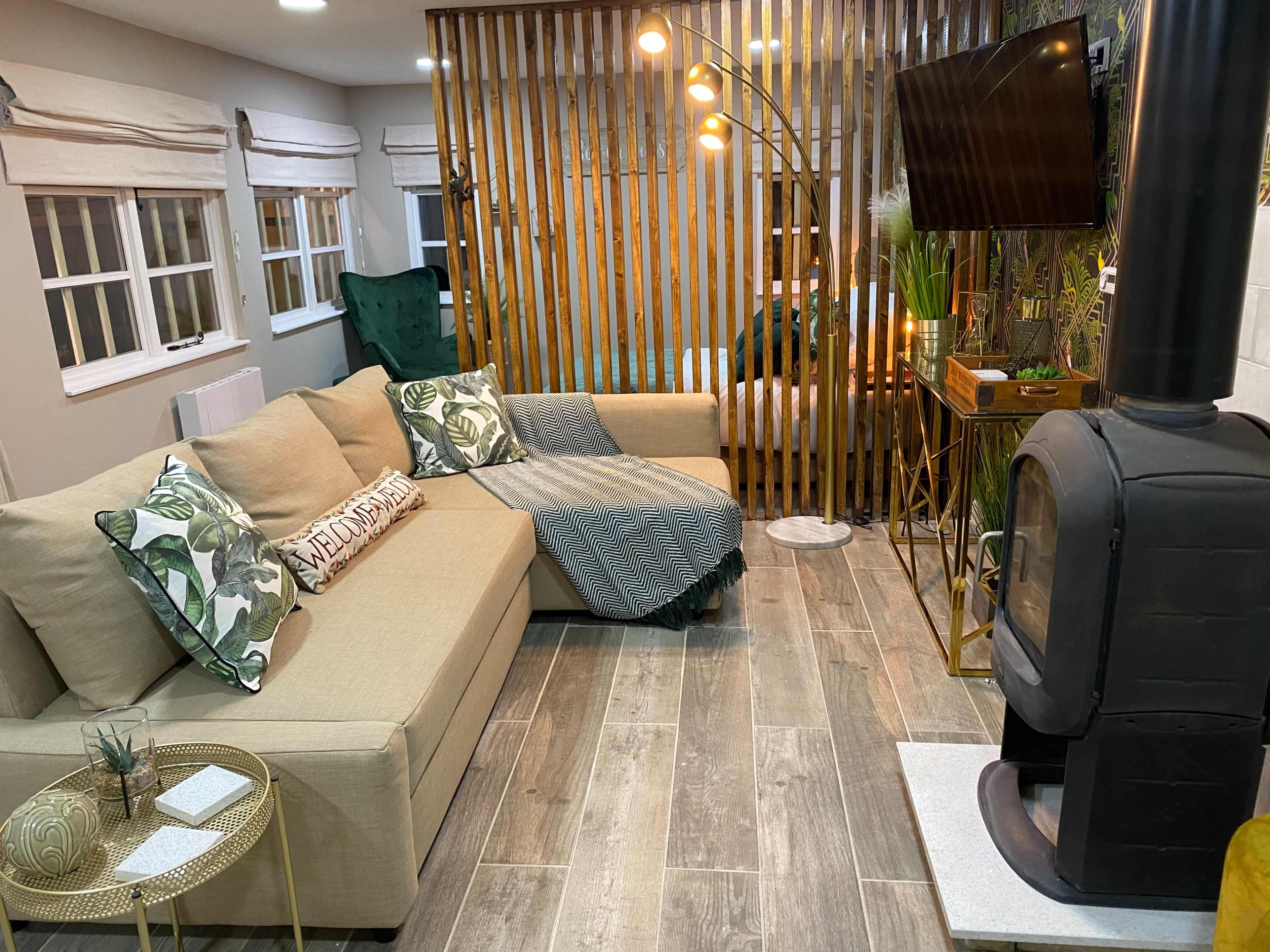 A cozy living area with a sectional sofa, a standing lamp, a television mounted on a wooden divider, and a modern wood stove.