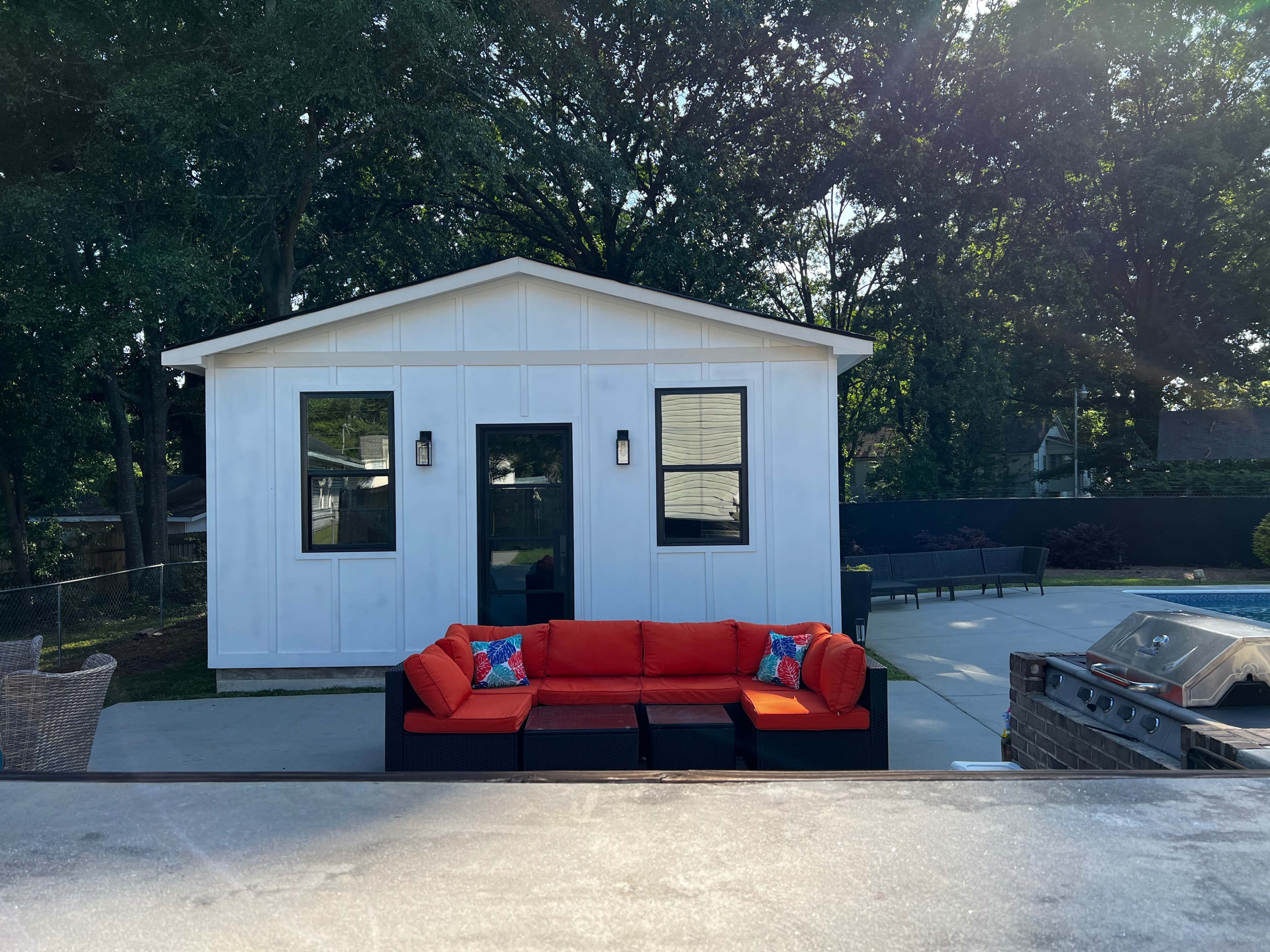 A white shed with large windows is situated behind an orange sectional sofa on a patio area surrounded by trees.